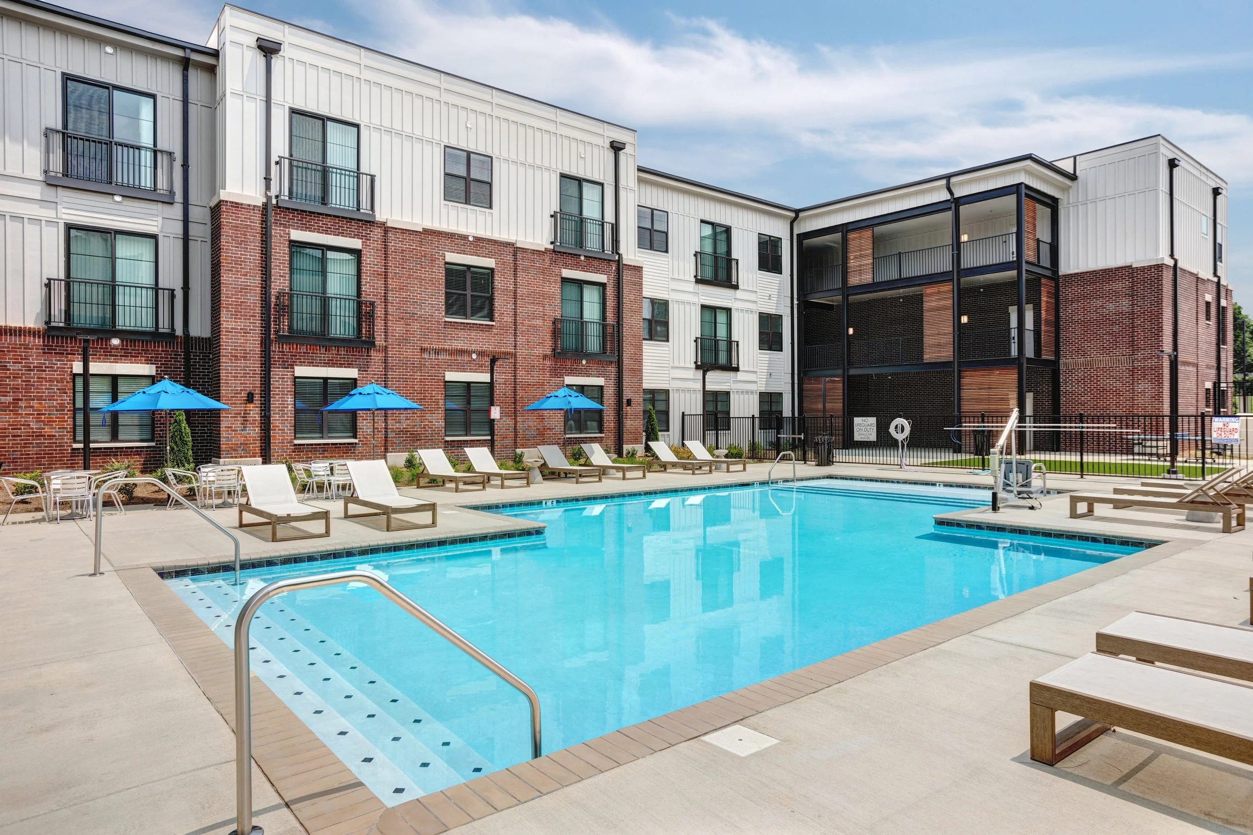 A swimming pool surrounded by lounge chairs, umbrellas, and residential buildings with multiple stories and balconies. The sky is partly cloudy.