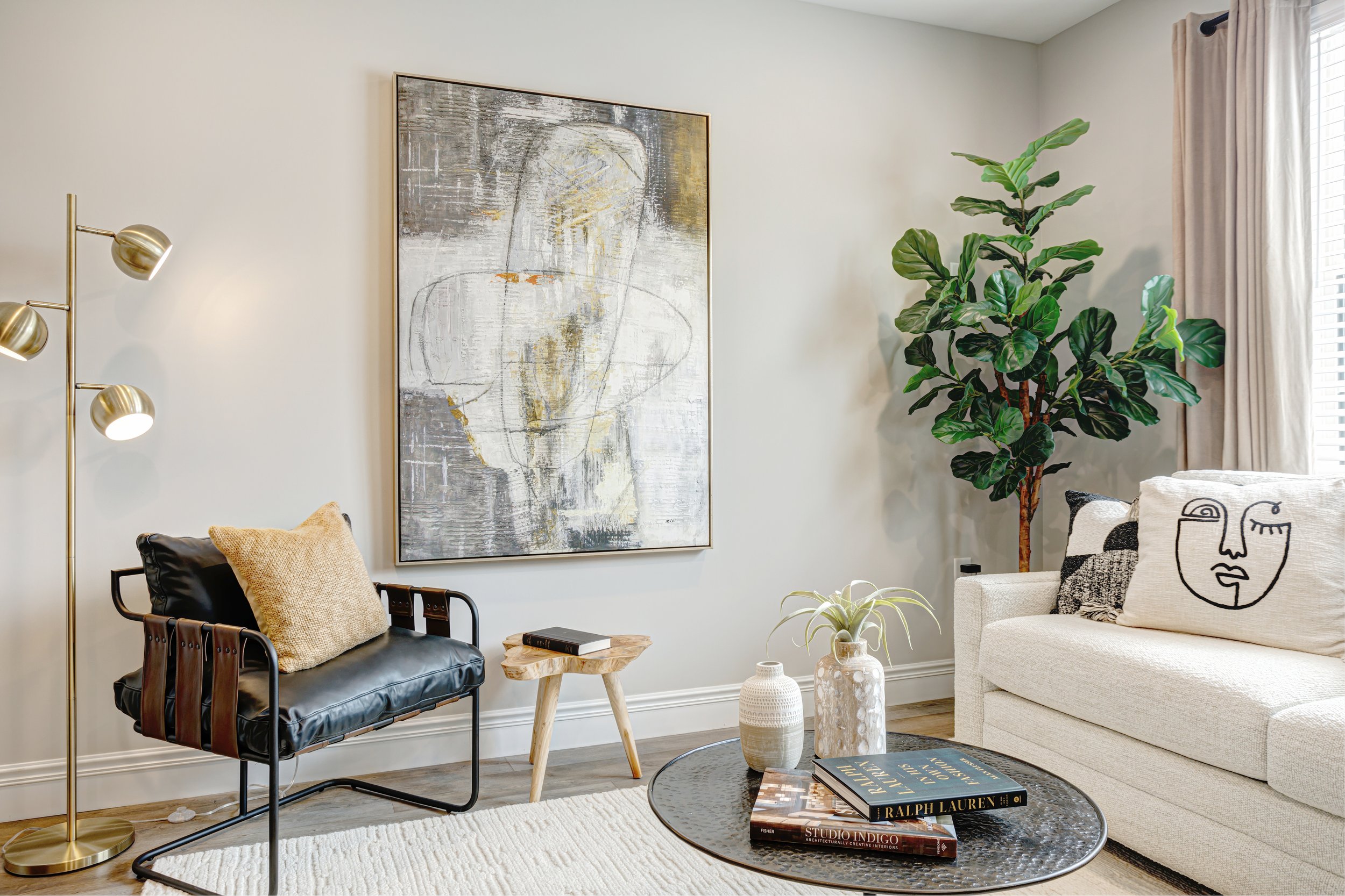 A modern living room corner with a black leather chair with a beige pillow, a small wooden side table, a tall gold floor lamp with three adjustable heads, a large abstract art print on the wall, a tall potted plant, a white upholstered sofa with blac