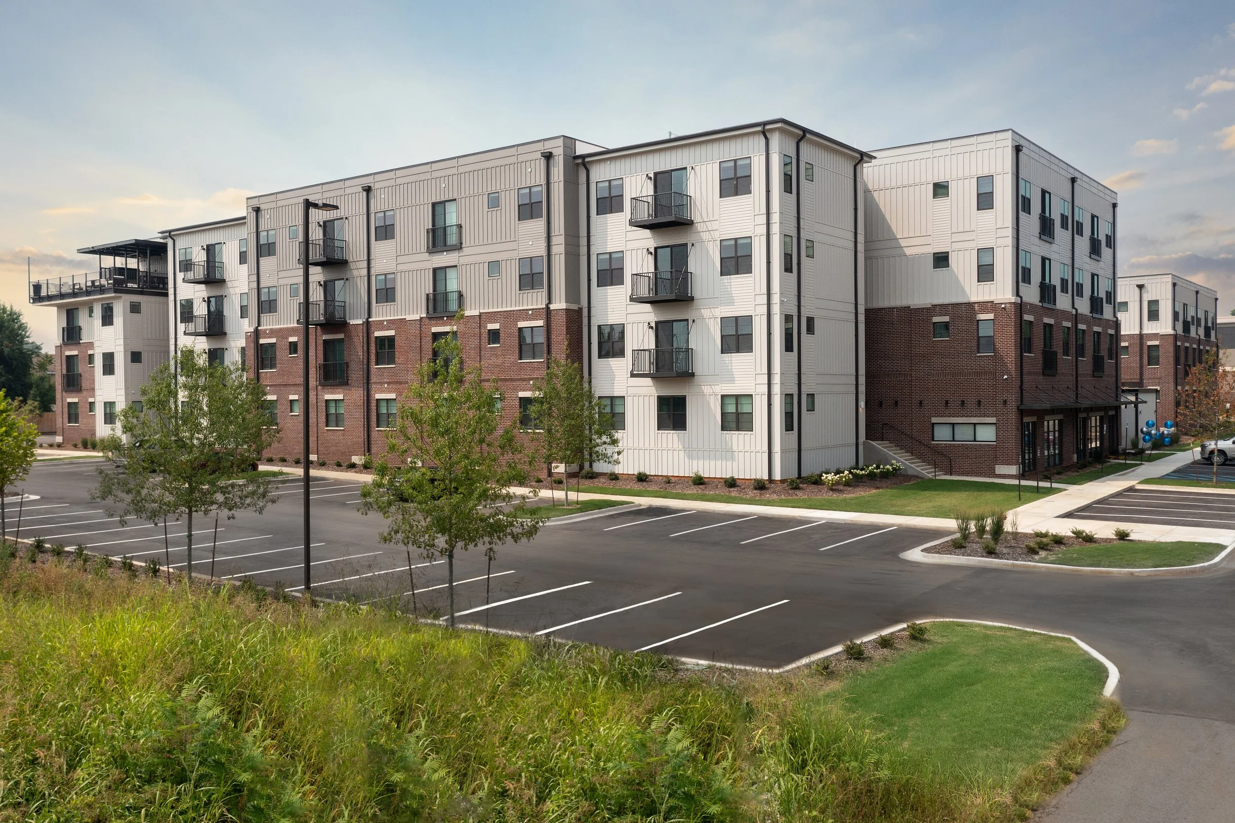 A multi-story apartment building with a brick and white siding exterior, multiple balconies, and a parking lot in front with trees and landscaped areas.