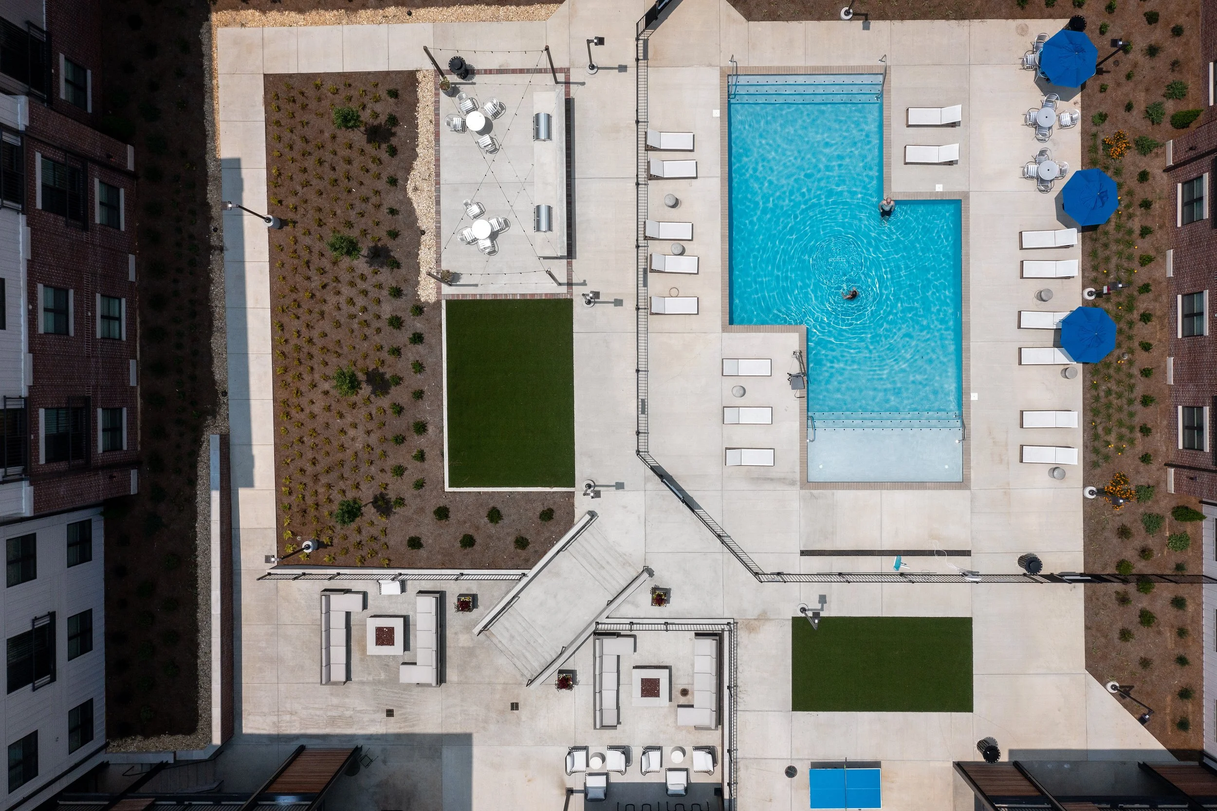 Aerial view of an apartment complex backyard with a swimming pool, lounge chairs, umbrellas, and landscaped areas.