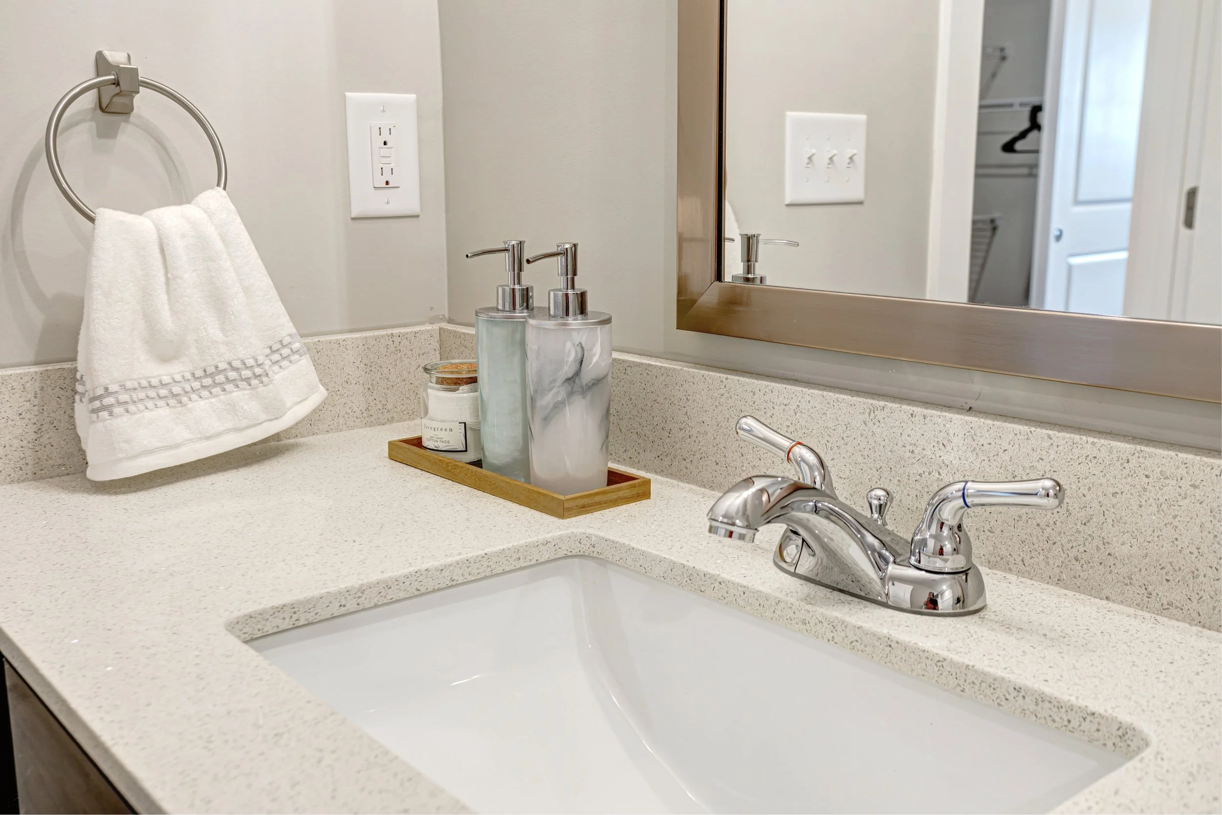 A bathroom sink with a bar of soap, two soap dispensers, and a candle on a wooden tray. A towel hangs on a metal ring, and a mirror is visible on the wall. An electrical outlet and a light switch are also in view.