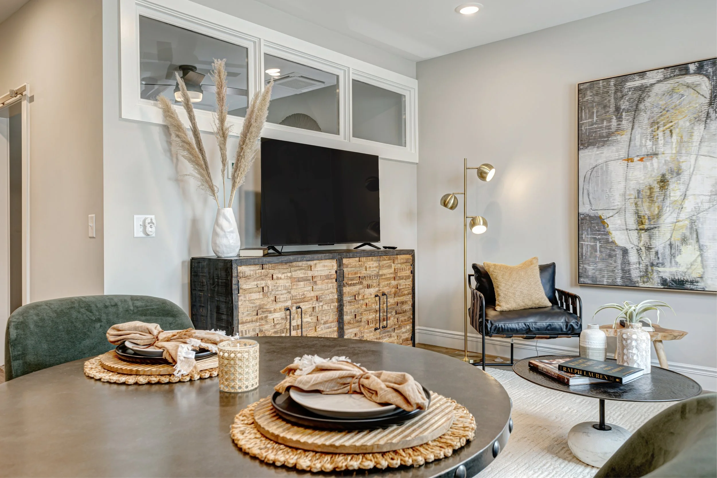 Living room with a black leather chair, a large abstract painting on the wall, a TV on a brick cabinet, a floor lamp with gold accents, a white vase with dried pampas grass, and a dining table with placemats, napkins, and bowls.
