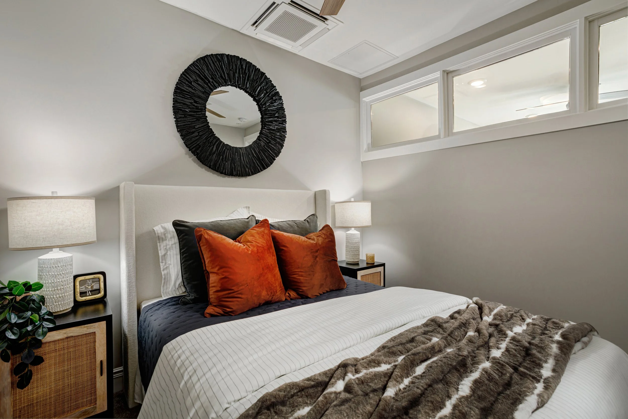 A bedroom with a white upholstered bed, black and orange throw pillows, a fur blanket, and a nightstand with a lamp, clock, and plant. There is a black circular mirror on the wall above the bed and a high horizontal window near the ceiling.