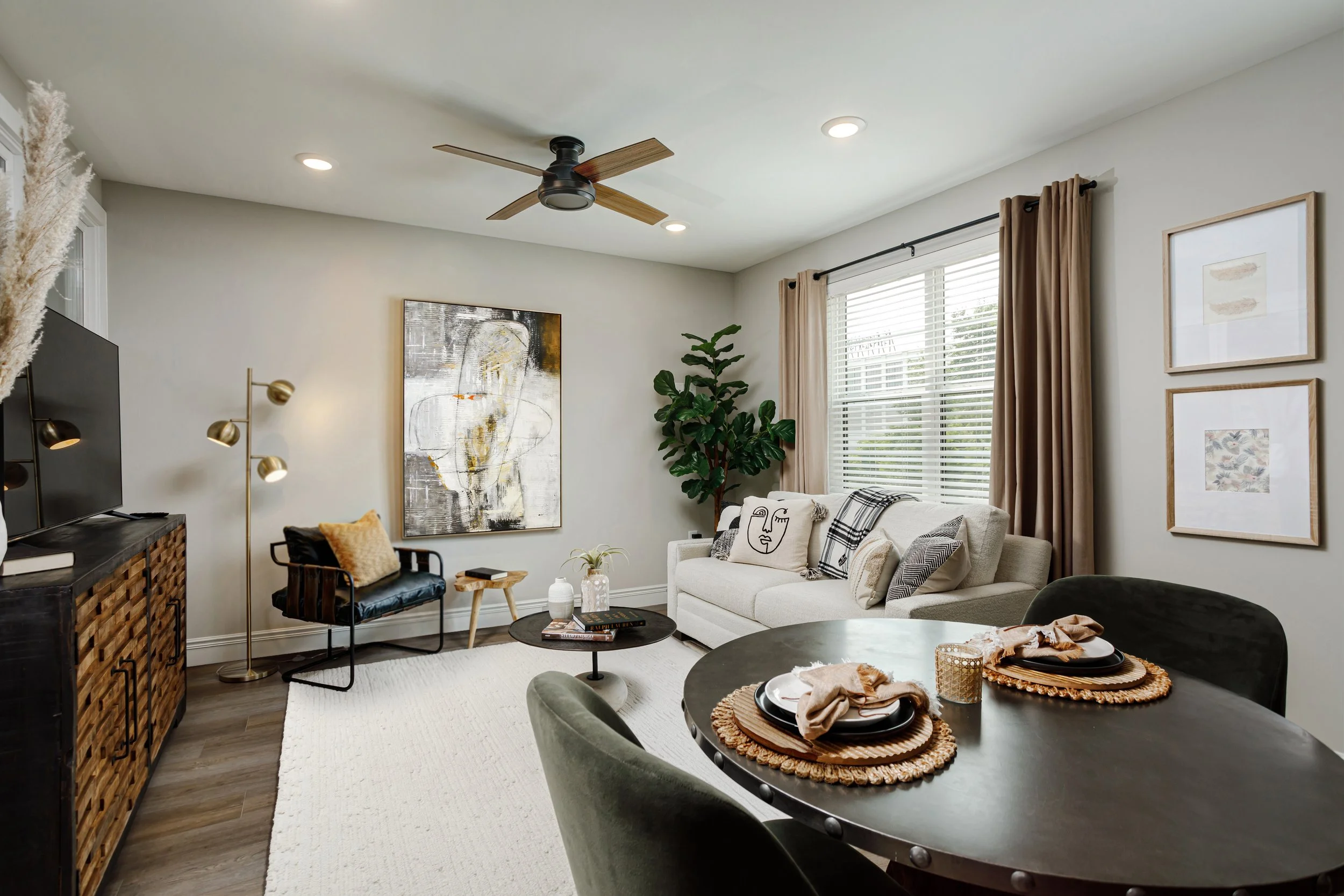 A modern living room with beige walls, a large window with beige curtains, a black chair with a yellow cushion, a white sofa with black and white pillows, a wooden coffee table, and wall art. There is a ceiling fan with wooden blades and recessed lig