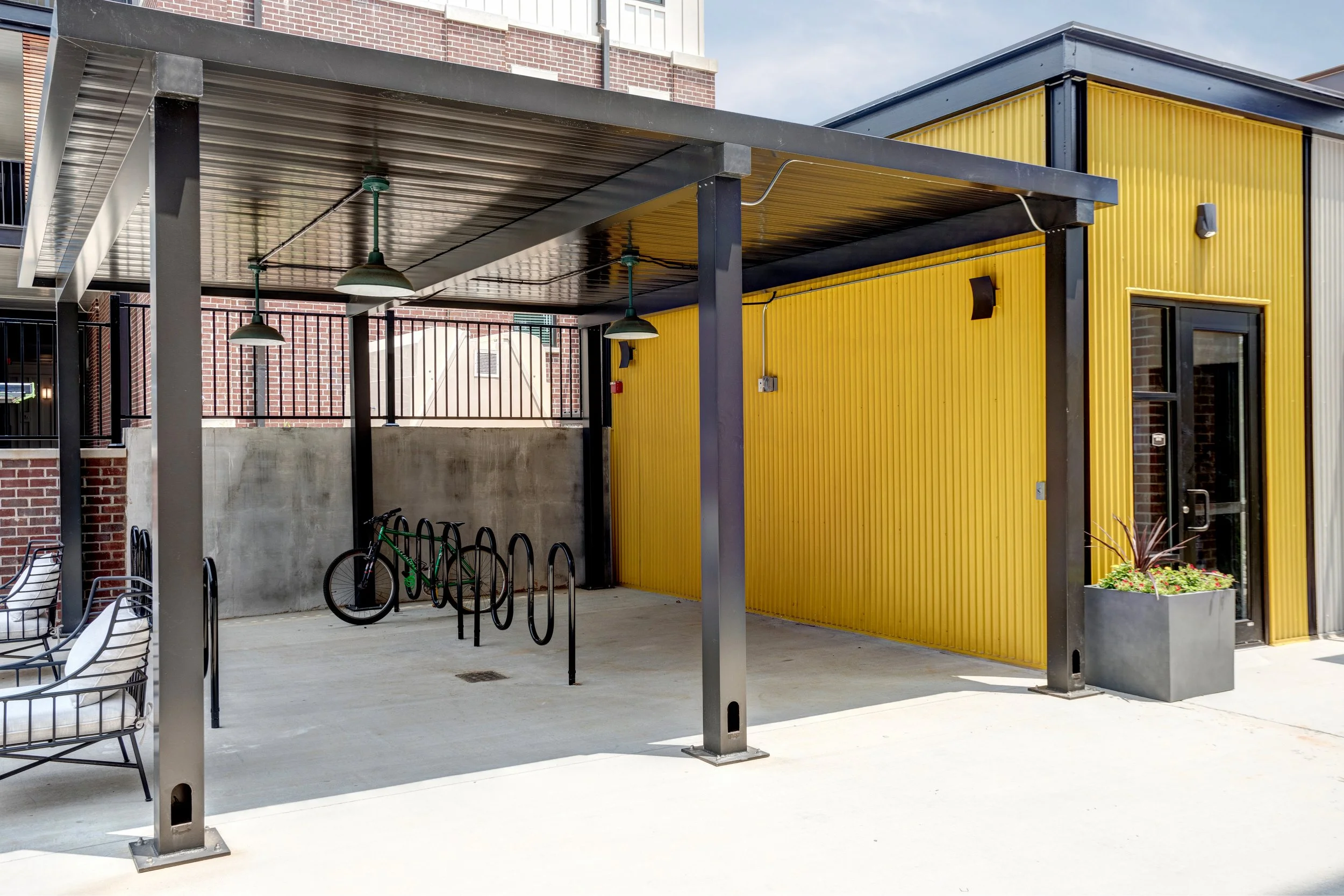 An outdoor seating area with metal chairs, a bike rack with two bikes, a yellow corrugated metal wall, and a covered structure with industrial-style hanging lights. There are brick buildings in the background and the sky is partly cloudy.