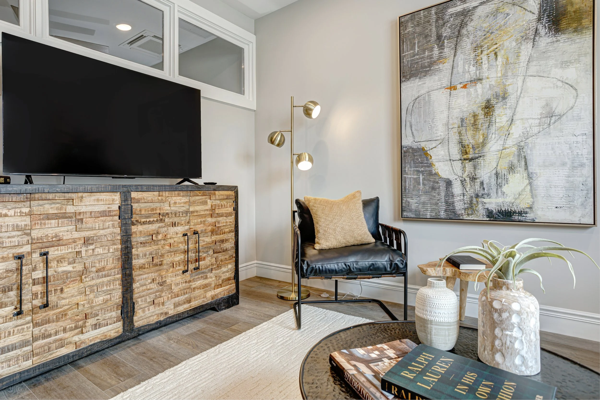 A living room corner featuring a flat-screen TV on a rustic wooden media console, a black armchair with a beige pillow, a round side table with decorative vases, a large abstract painting on the wall, a gold-colored floor lamp, and a cream-colored ru