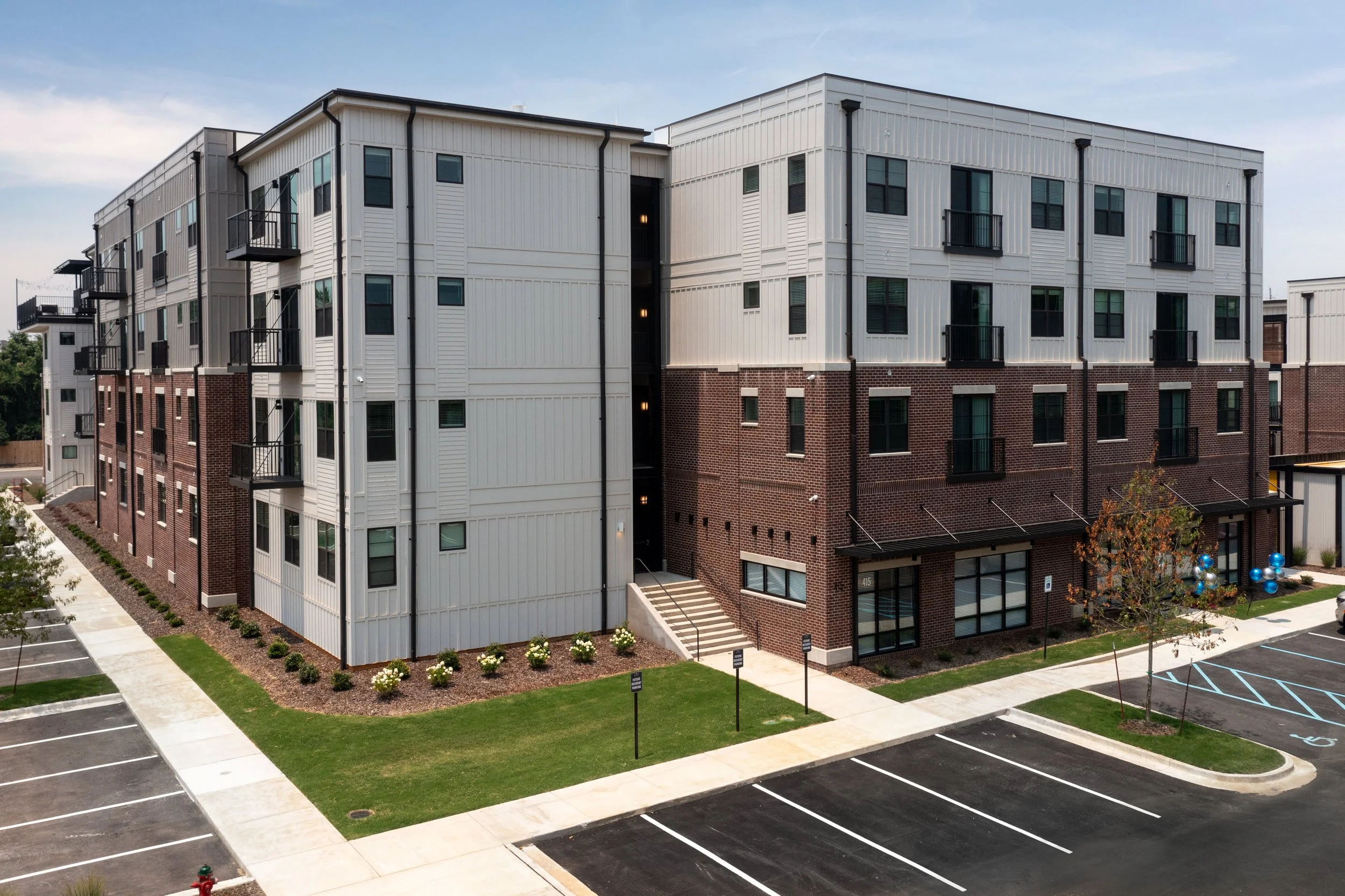 Image of a modern multi-story residential building with a mix of brick and white siding exterior, surrounded by a parking lot, sidewalks, and landscaped areas with small bushes and a tree. The sky is partly cloudy.