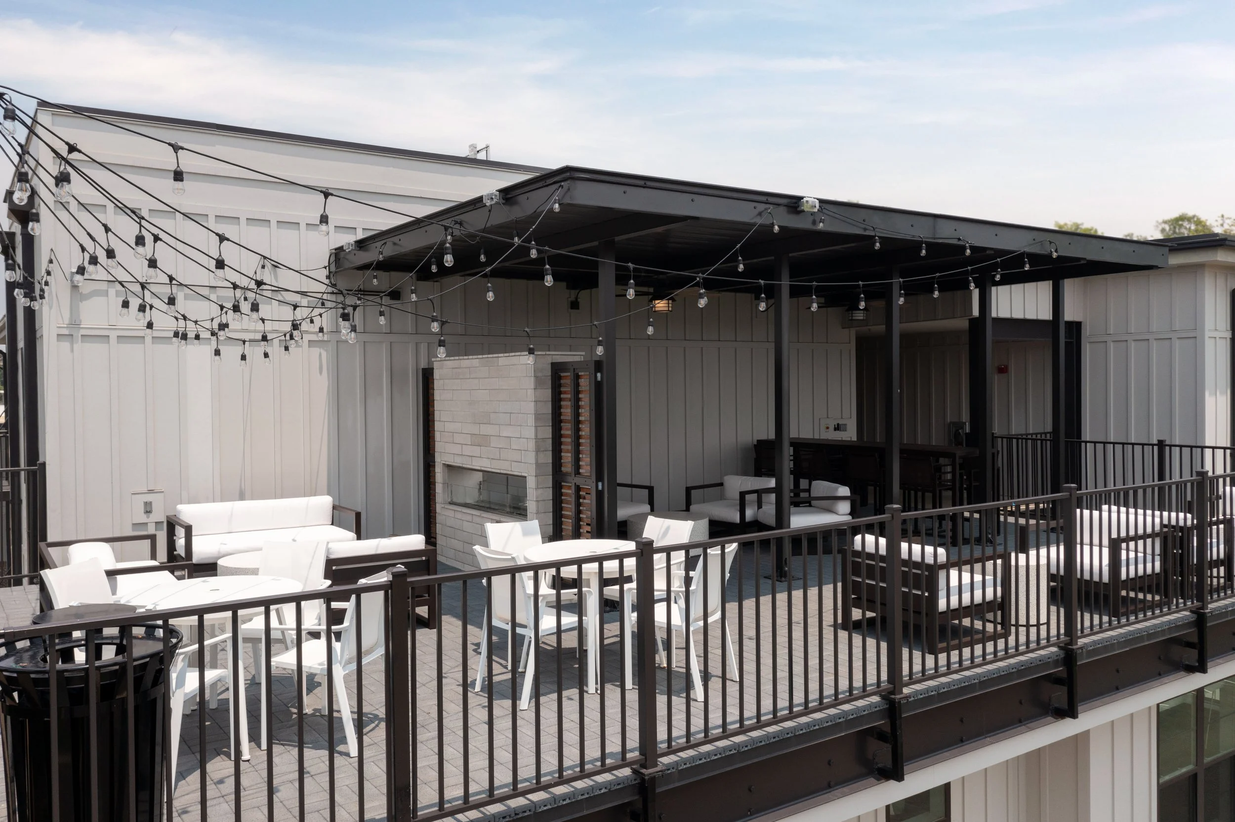 Outdoor balcony with white chairs, black railing, string lights, and an under-the-roof seating area with chairs and a fireplace.