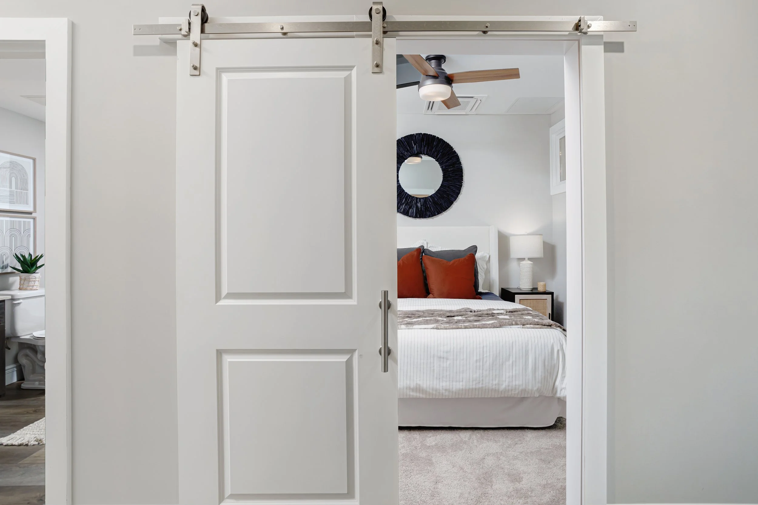 A white sliding barn door partially open, revealing a bedroom with bed, dark blue circular wall mirror, and window.