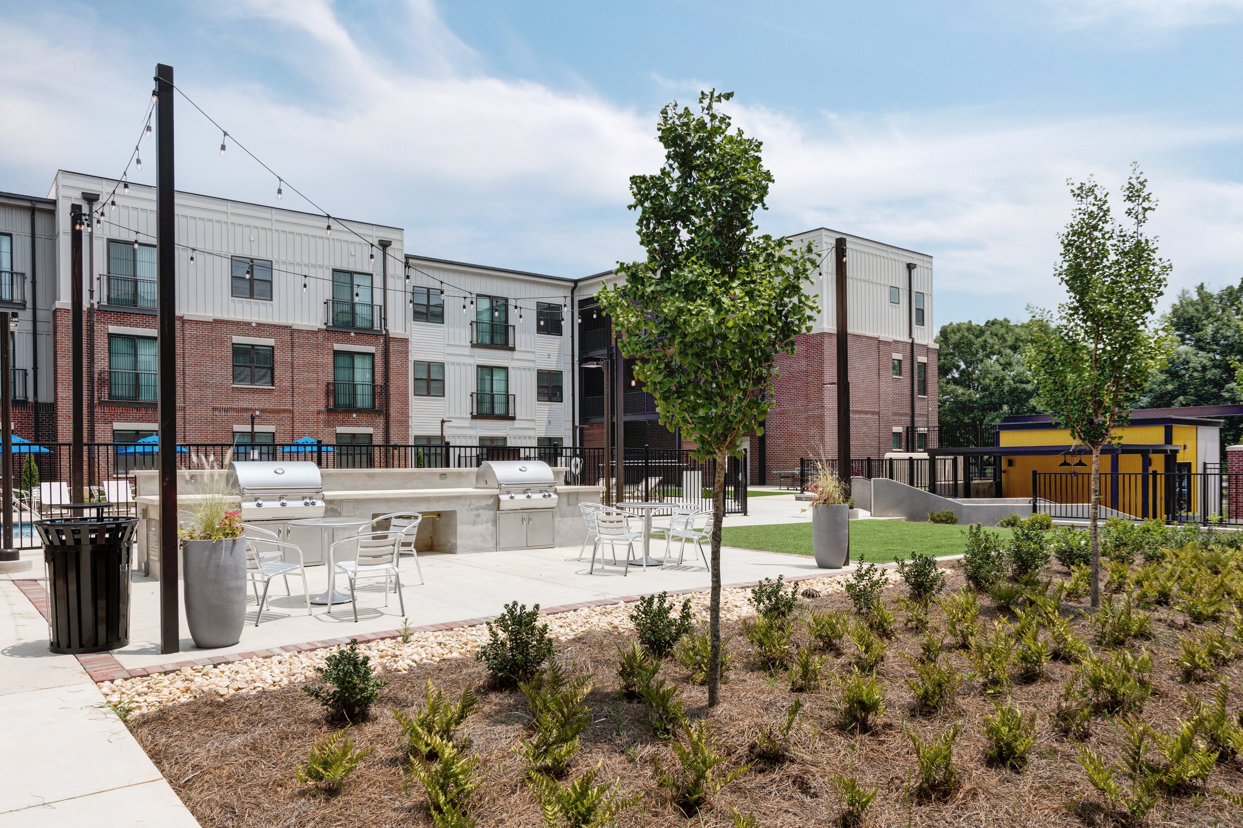Outdoor communal area with trees, potted plants, white chairs, and grills, surrounded by a fence and apartment buildings in the background under a blue sky.