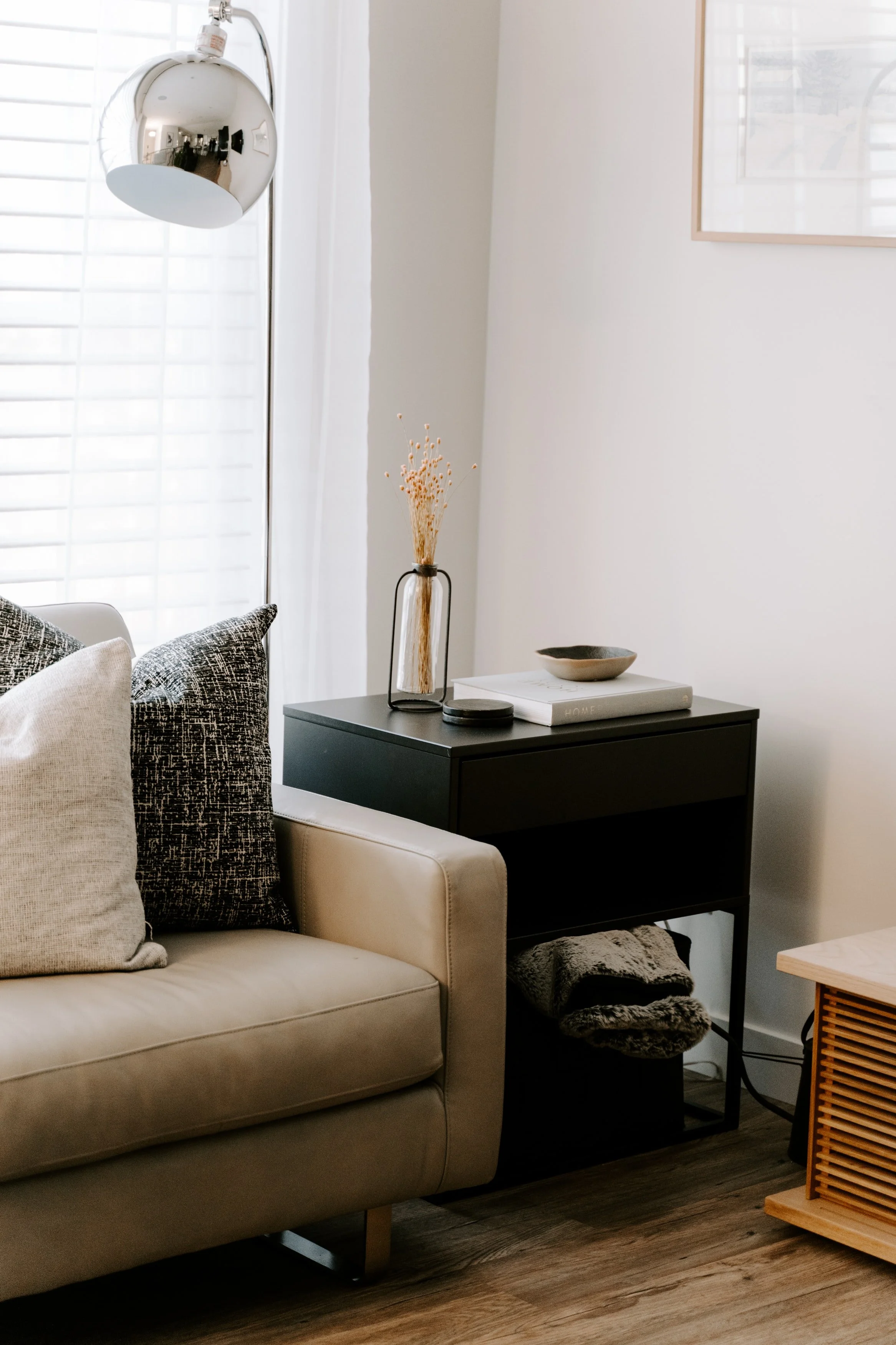 A living room corner with a beige sofa, black and patterned throw pillows, a black side table with a decorative vase, and a small side table with a wooden slatted door. There is a framed picture on the white wall and a window with white blinds.