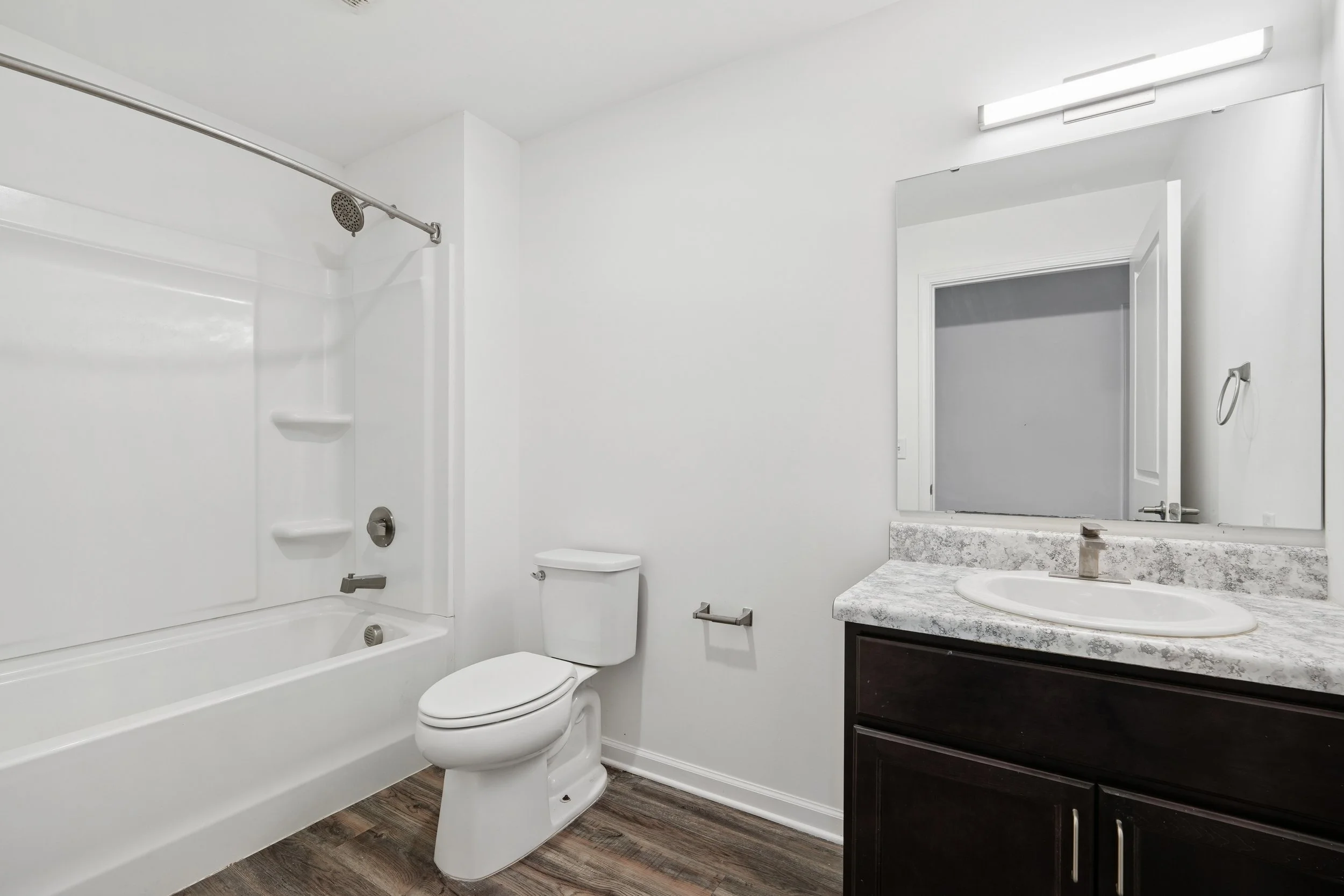 A small modern bathroom with a bathtub and shower, toilet, and a vanity with a sink and mirror. The bathroom has white walls, a grey marble countertop, and dark cabinetry. There is a towel ring next to the mirror, a shower curtain rod, and wood-look flooring.