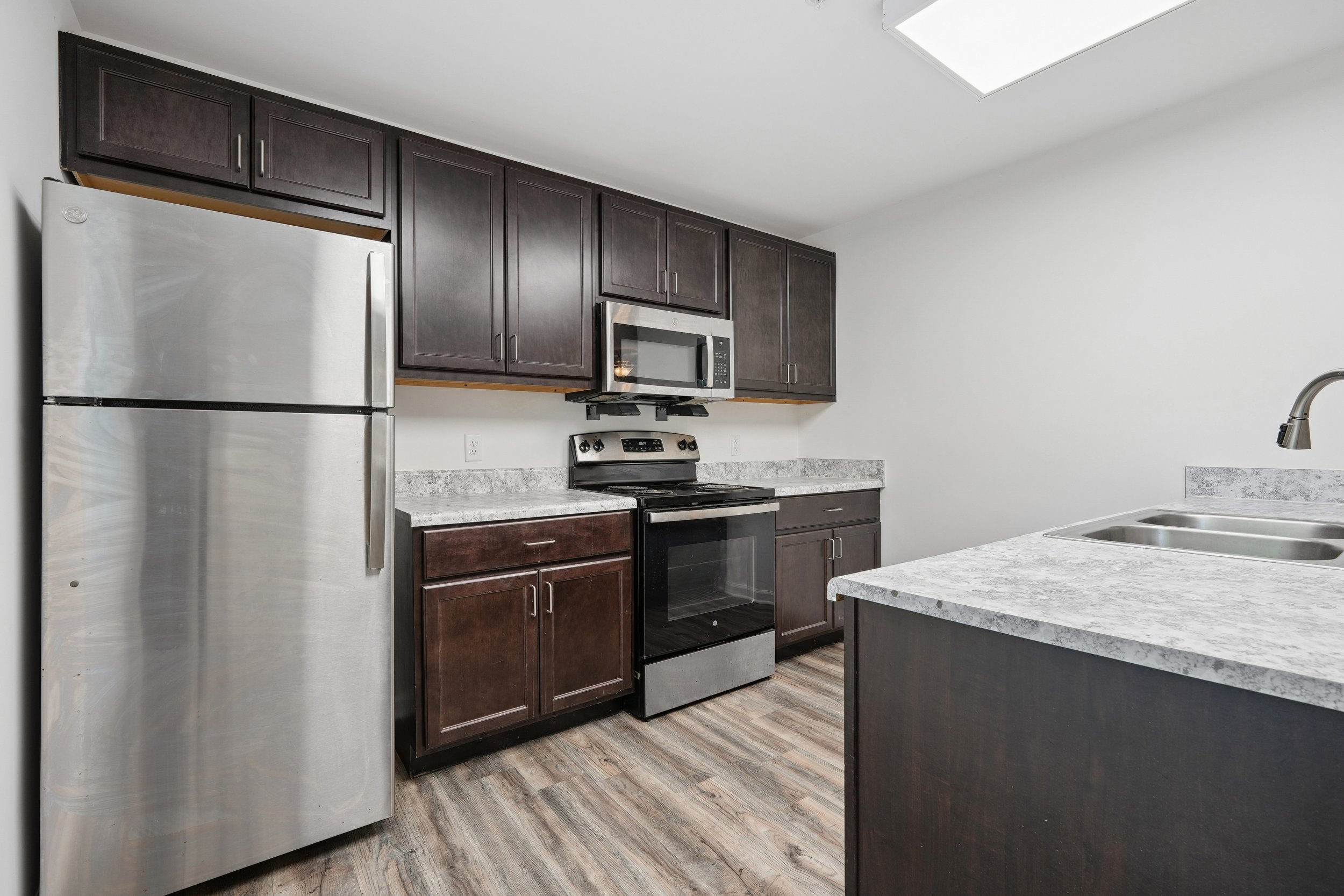 Kitchen with dark wood cabinets, stainless steel refrigerator, microwave, stove, and a gray and white speckled countertop. The floor is wood, and there is a white wall with a ceiling light.