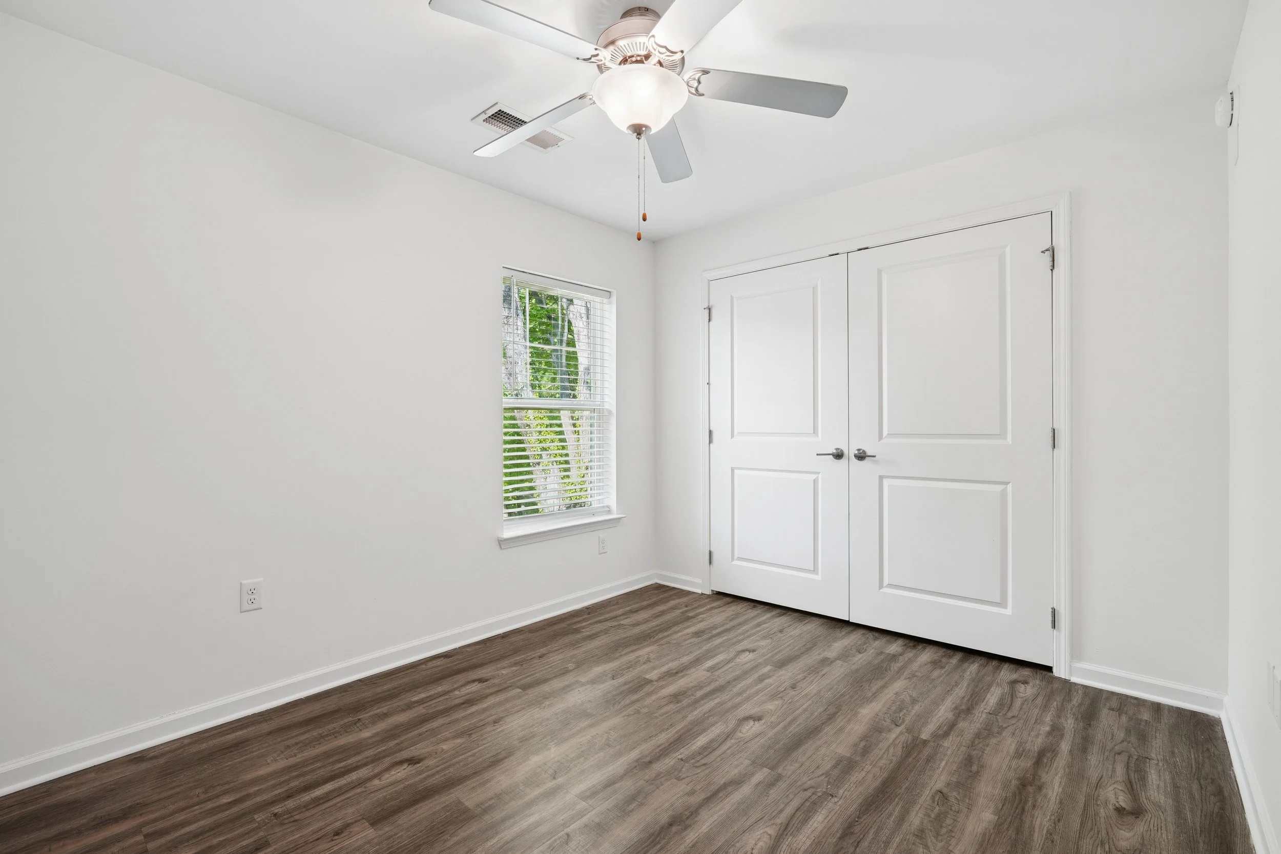 Empty white room with dark wood flooring, a ceiling fan, a window with blinds, and a large double closet with white doors.