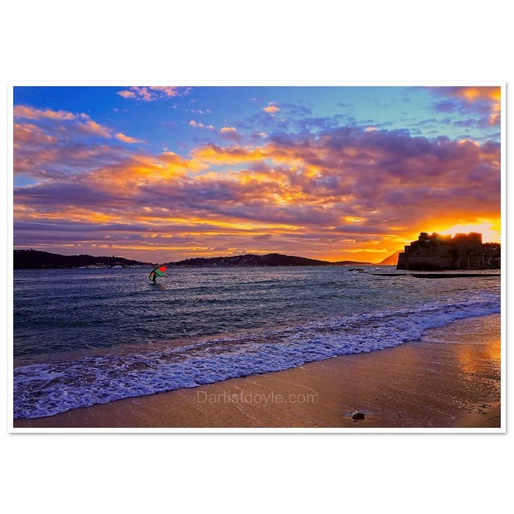 A sunset over the ocean with a colorful sky, a kite surfer in the water, and a rocky cliff on the right side.