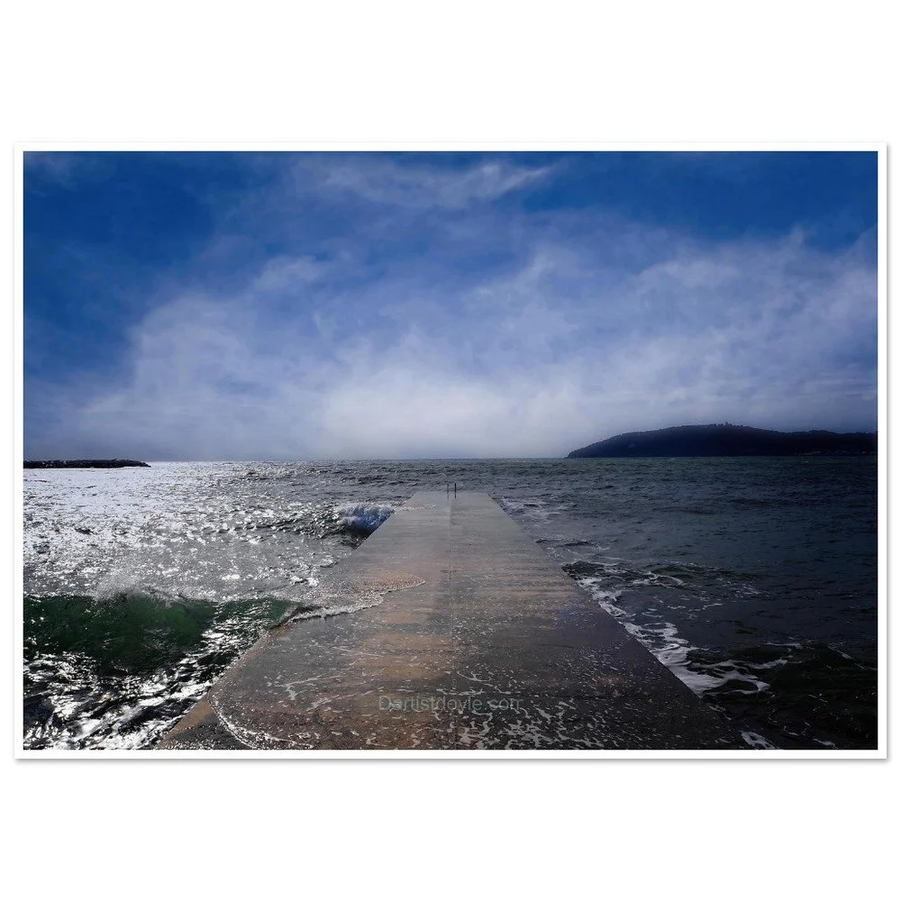A concrete pier extends into the ocean with waves washing over it, under a partly cloudy sky with a landmass in the distance.