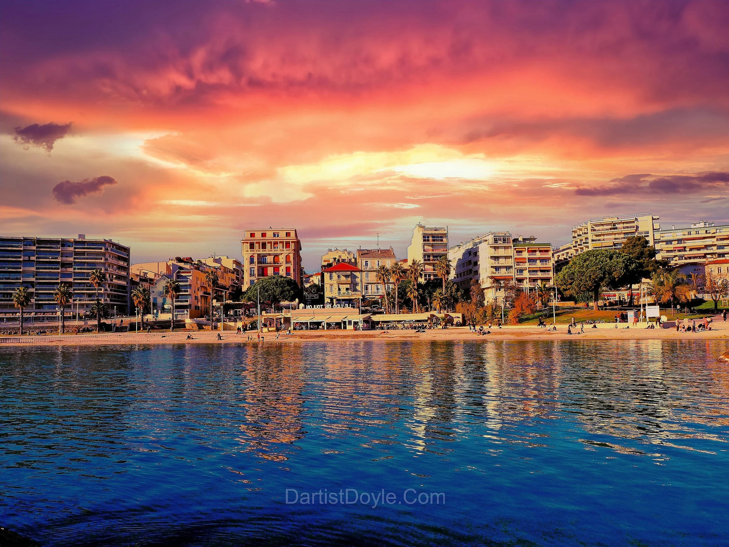 A cityscape during sunset with colorful clouds in the sky and buildings along the waterfront, with reflections visible on the water surface.