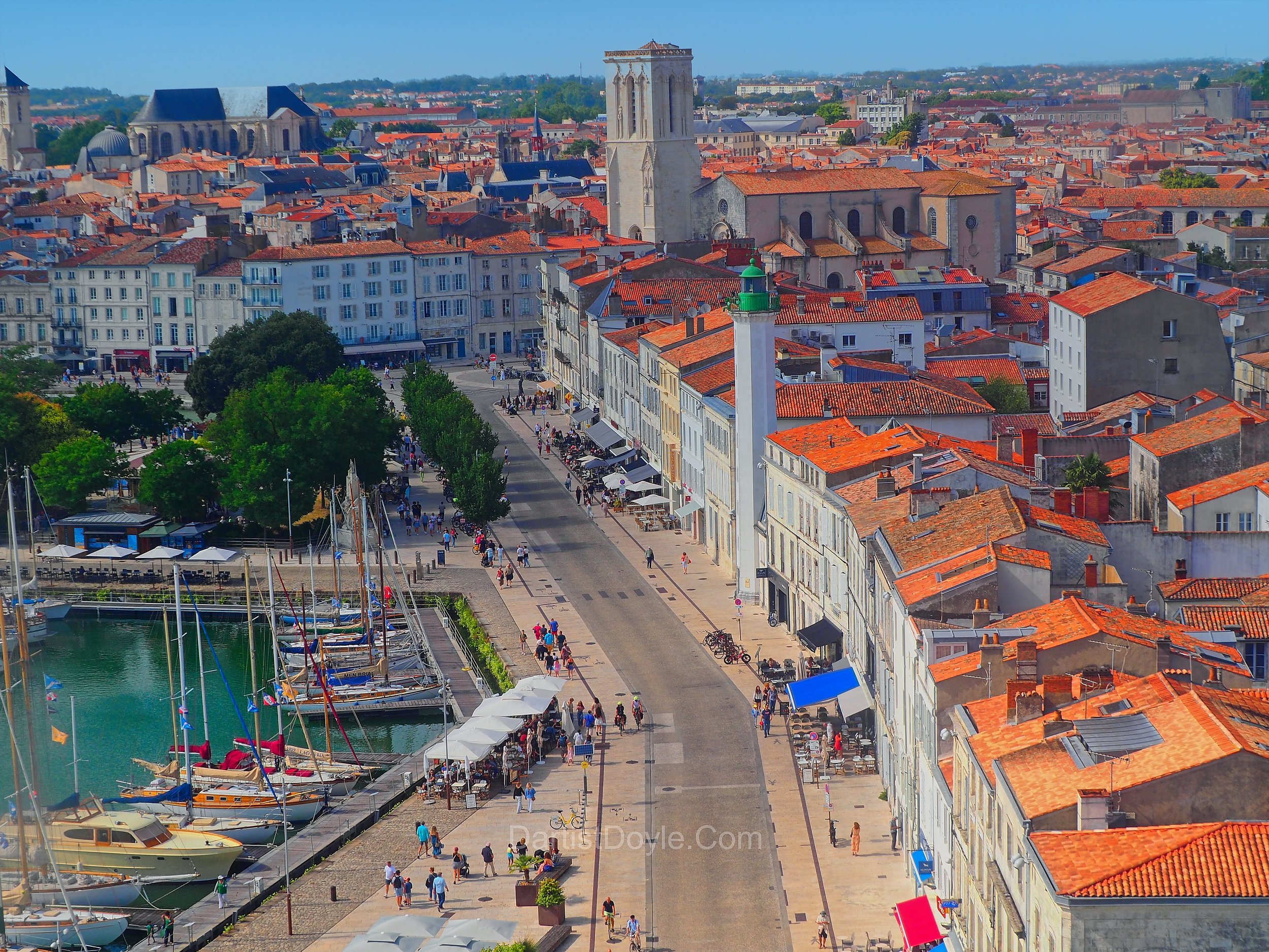Aerial view of a coastal European city with red-tiled roofs, historic churches, a marina with sailboats, and people walking along the waterfront promenade.