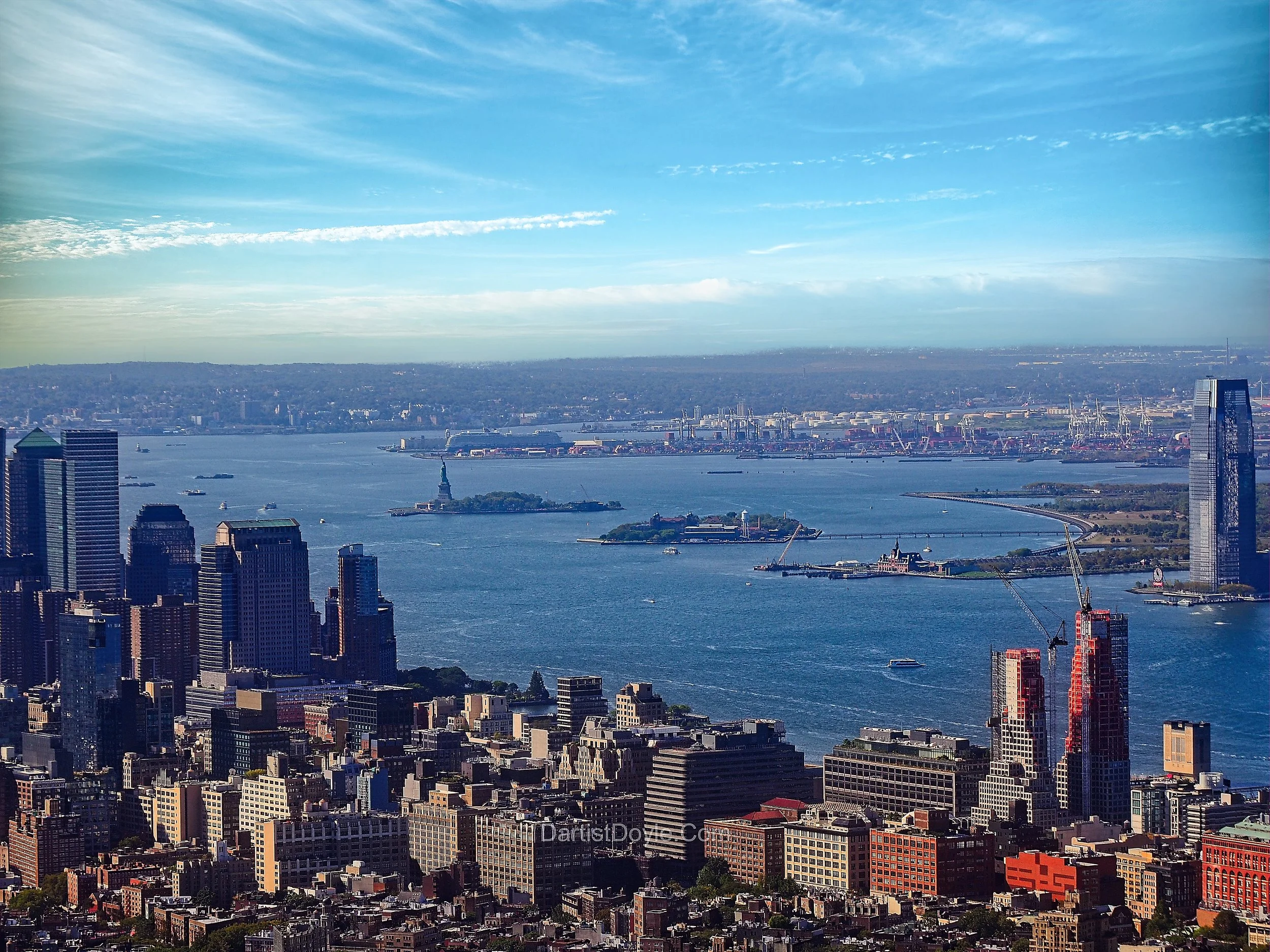 A panoramic view of New York City skyline with tall buildings in the foreground, a body of water with boats, and an island with the Statue of Liberty, under a partly cloudy sky.