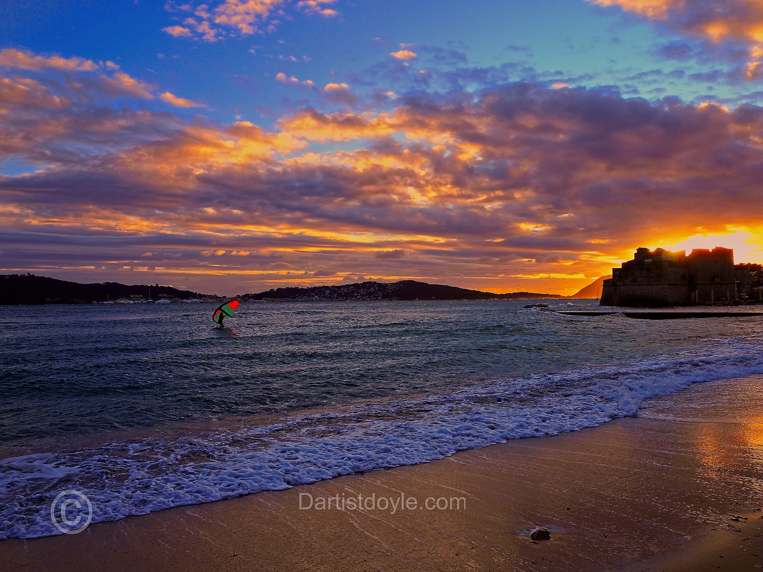 A sunset over a body of water with a city or fort silhouette on the right side and a person kite surfing in the water on the left side. The sky is filled with colorful clouds illuminated by the setting sun.