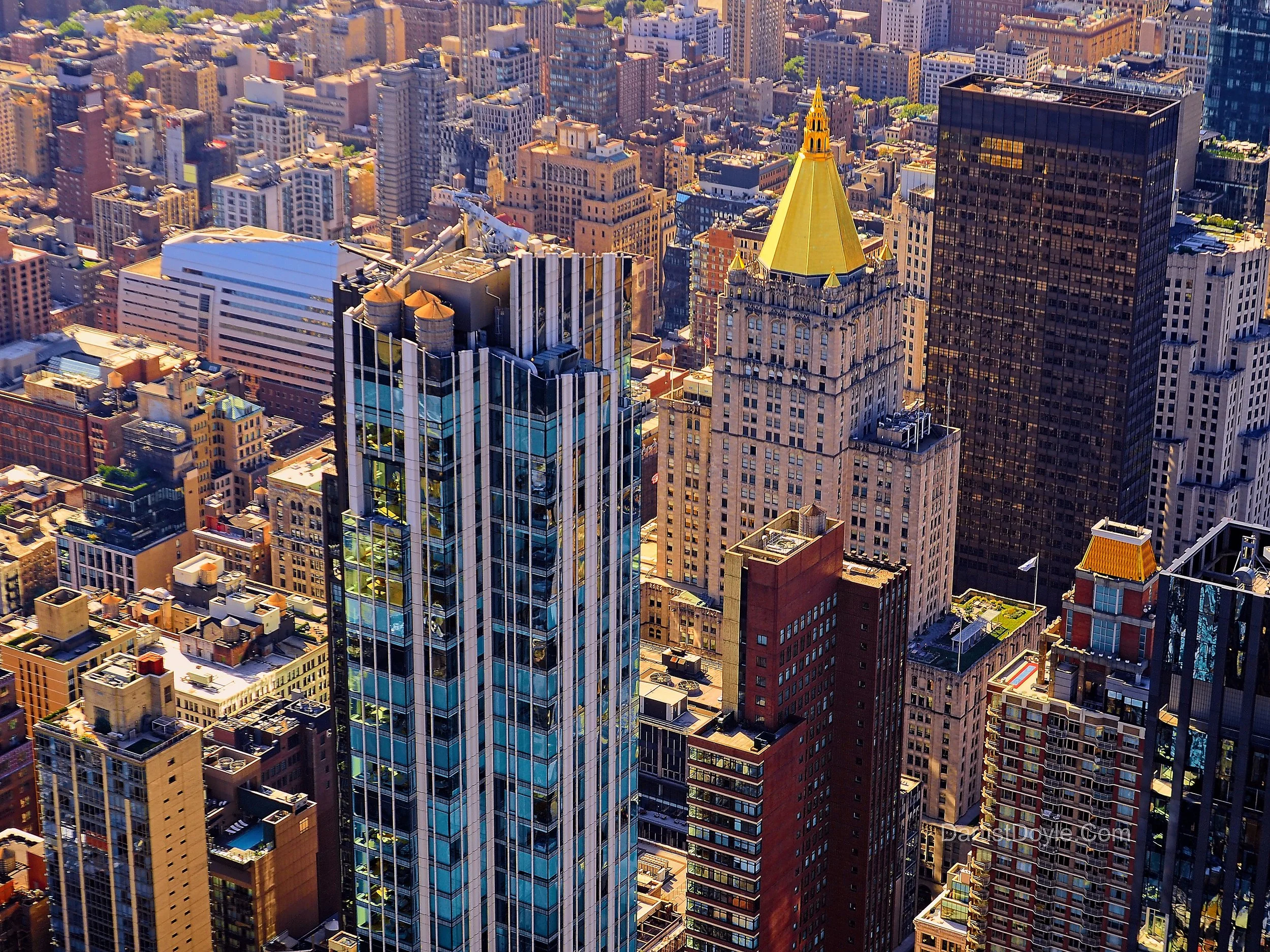 Aerial view of a dense cityscape with numerous tall buildings, including a prominent skyscraper with a gold spire, surrounded by various office and residential buildings.