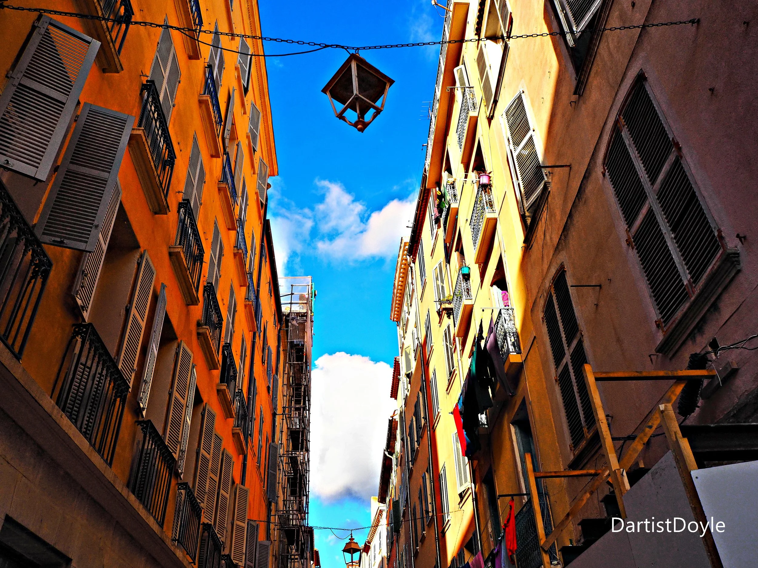 View of colorful apartment buildings with open window shutters and laundry hanging on balconies, under a bright blue sky with some clouds.