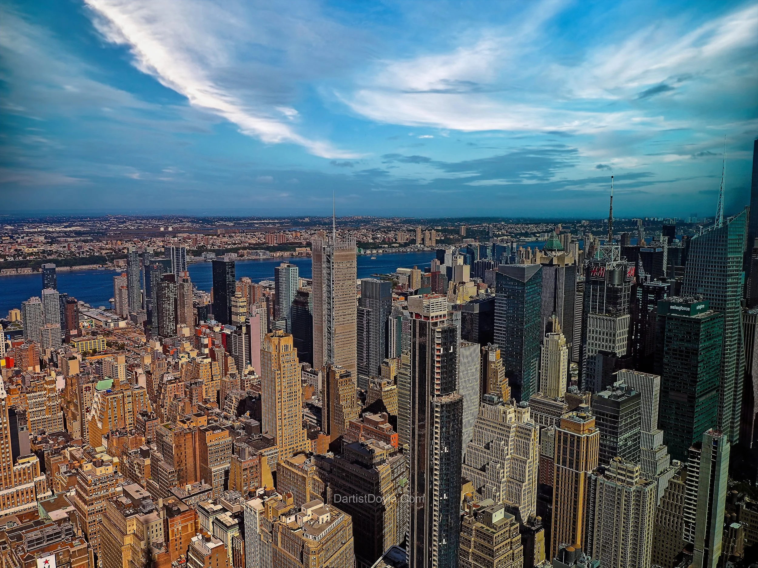 A panoramic view of New York City skyline with tall skyscrapers, including the Empire State Building, overlooking the Hudson River under partly cloudy skies during daytime.