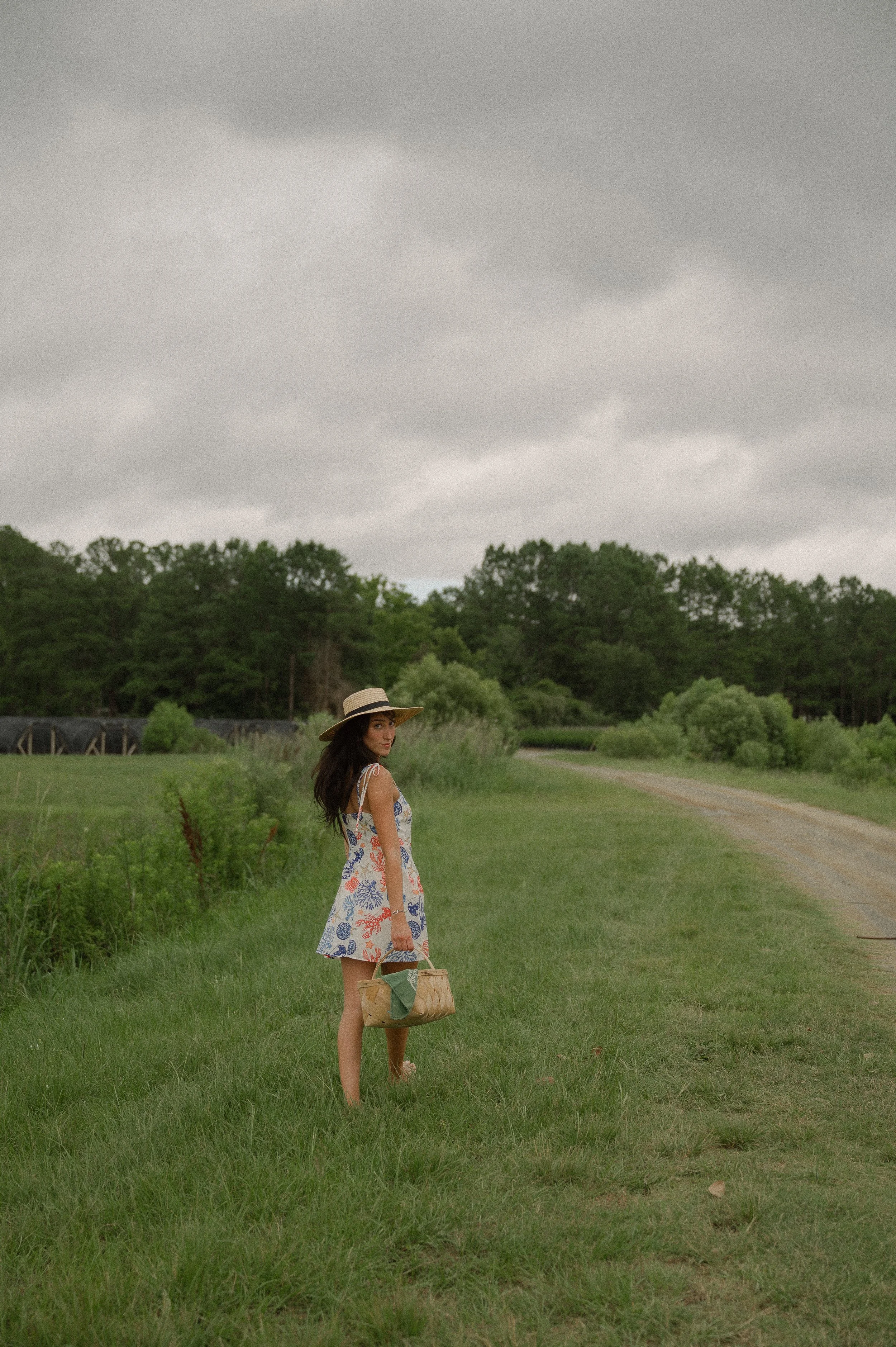 A woman in a colorful summer dress and wide-brimmed hat standing barefoot on a grassy field, holding a woven bag, with trees and a cloudy sky in the background.