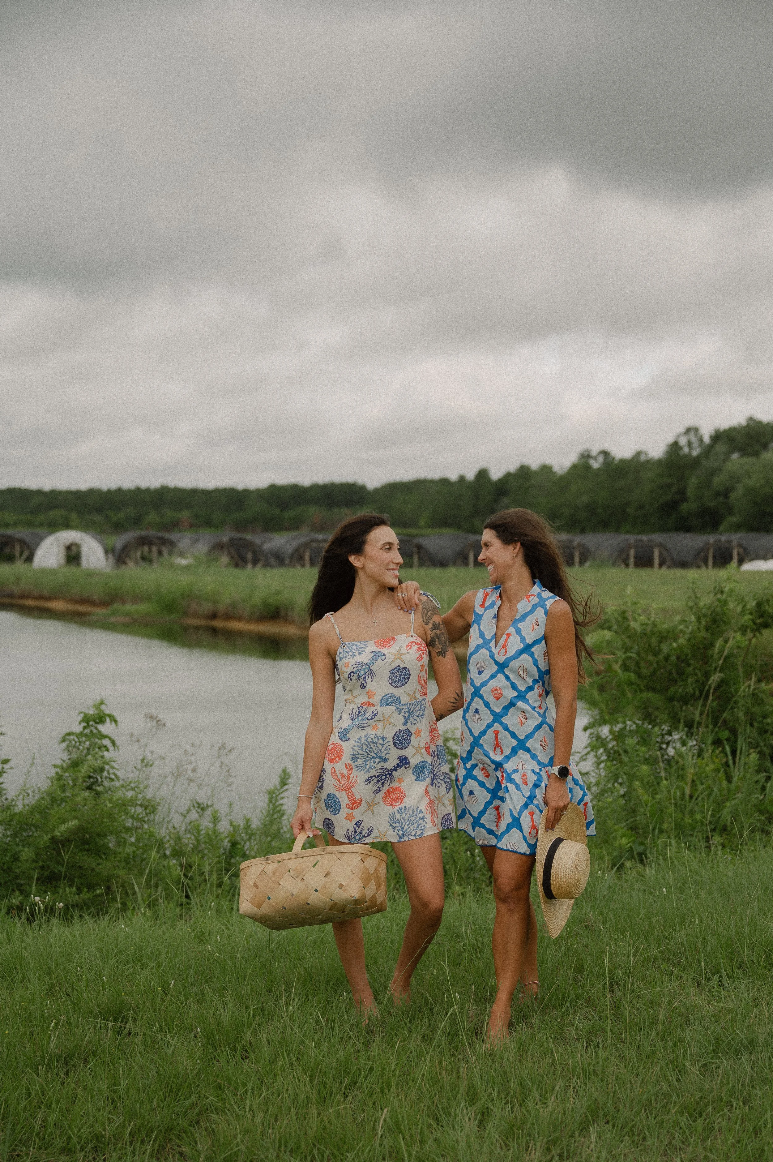 Two women walking on grass by a river under a cloudy sky, carrying a basket and a hat, and smiling at each other