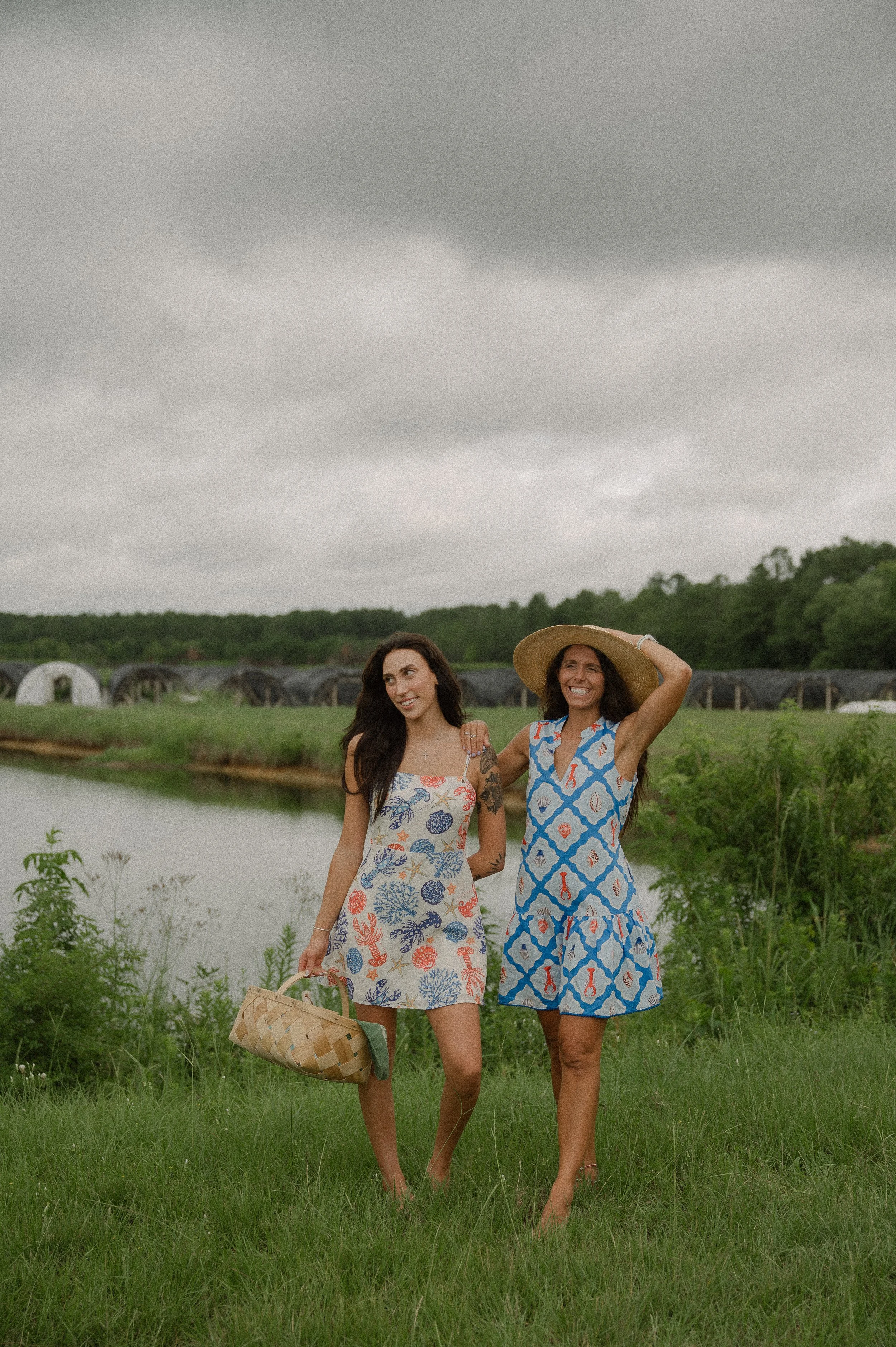 Two women walking outdoors in a grassy area near a body of water, with a cloudy sky and trees in the background. One woman is wearing a sleeveless dress with a colorful floral pattern and holding a woven basket; the other woman is wearing a sleeveless dress with a blue and orange pattern and a large sunhat, smiling and talking.