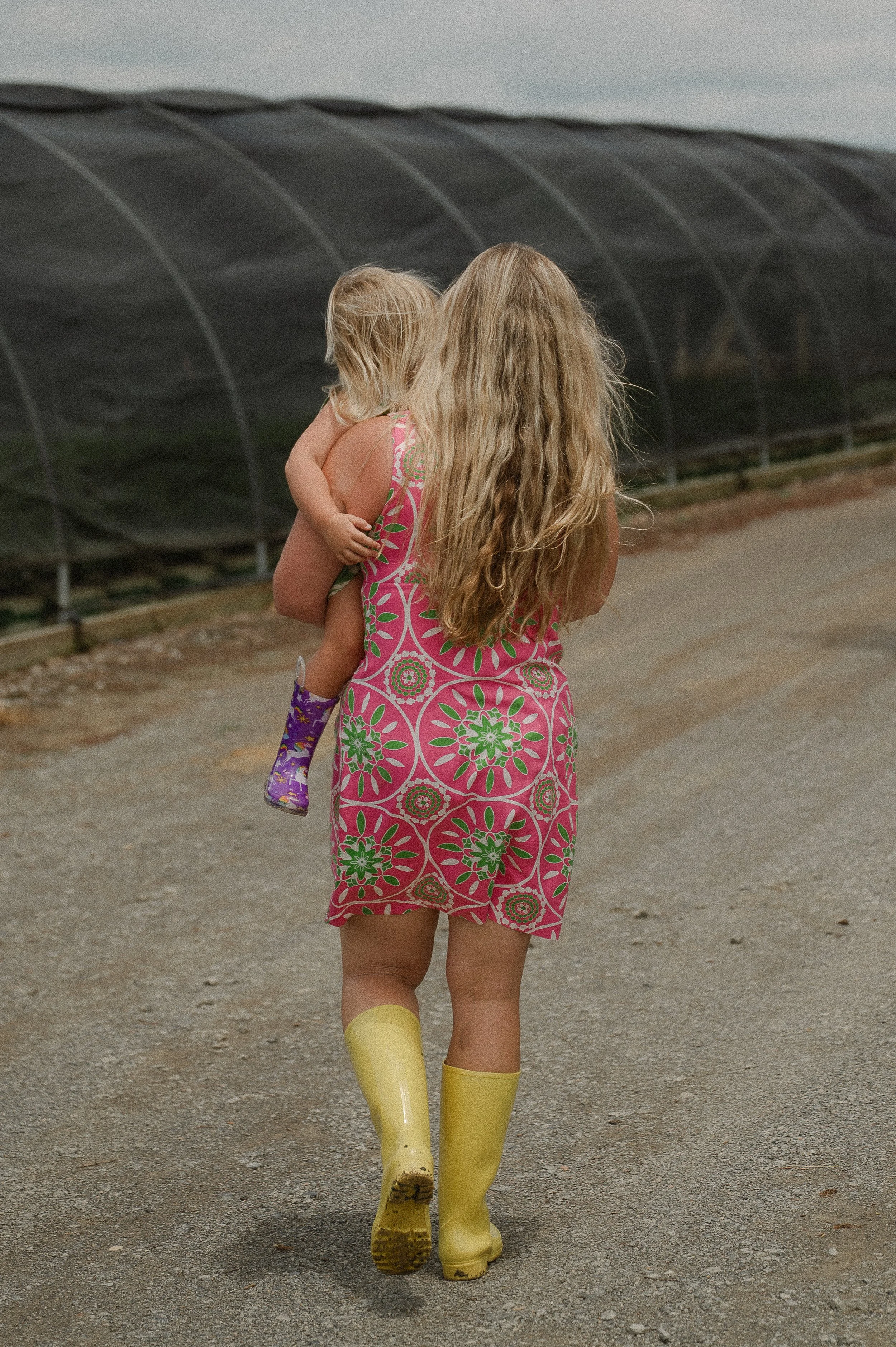 A woman with long, wavy blonde hair, wearing a pink sleeveless dress with a green and white floral pattern, yellow rubber rain boots, and carrying a small child with blonde hair on her back, walking on a dirt path near what appears to be a greenhouse structure.