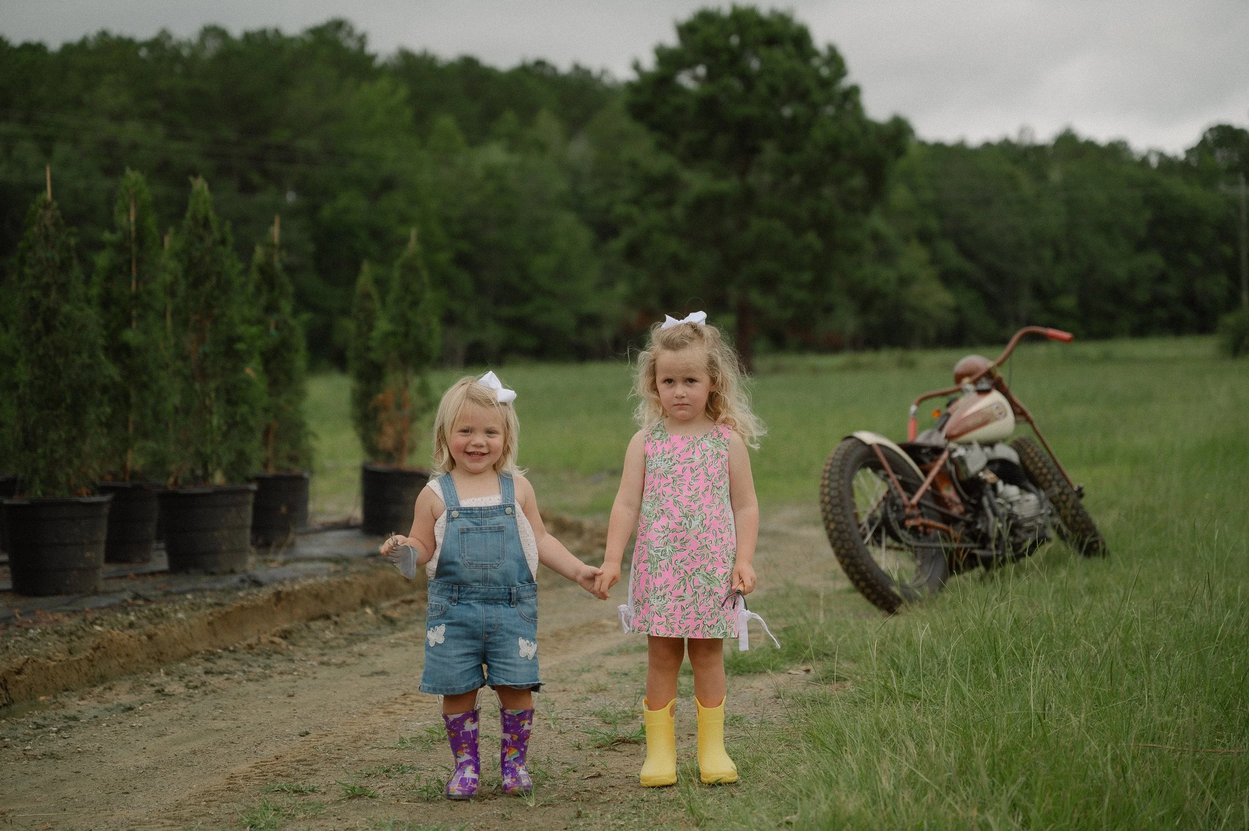 Two young girls holding hands while walking outdoors on a cloudy day. One girl has blonde curly hair, wearing a pink floral dress and yellow rain boots. The other girl has straight blonde hair with a white bow, wearing denim overalls, a white shirt, and colorful rain boots. In the background, there are potted trees, a dirt path, and a vintage motorcycle.