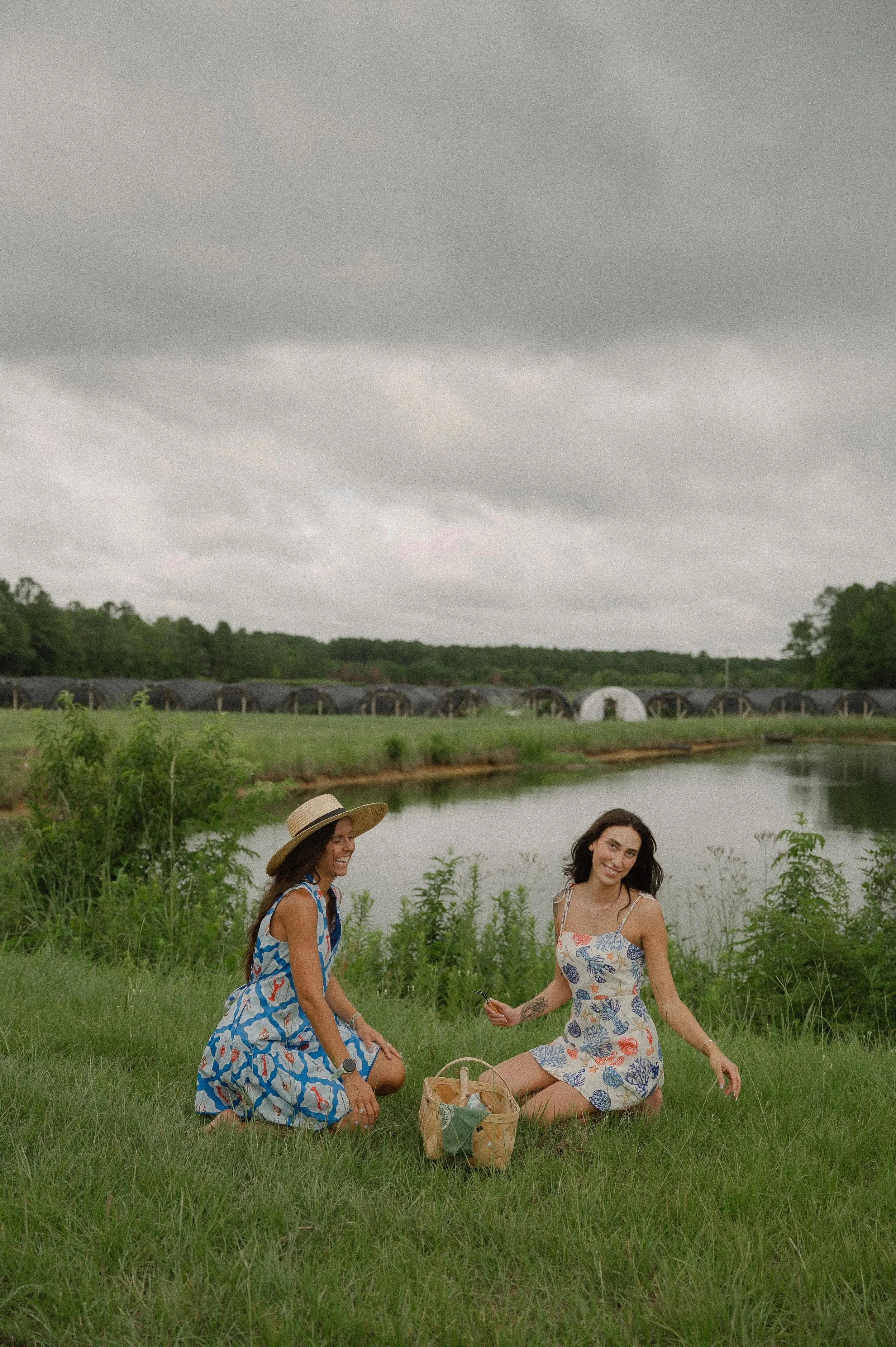 Two women sitting on the grass near a lake, smiling and relaxing on a cloudy day. One wearing a wide-brimmed hat and a blue and red patterned dress, the other with dark hair, a floral dress, and tattoos, holding a phone. A picnic basket is nearby, with lush greenery and trees in the background, and a farm with greenhouses and a pond under a cloudy sky.