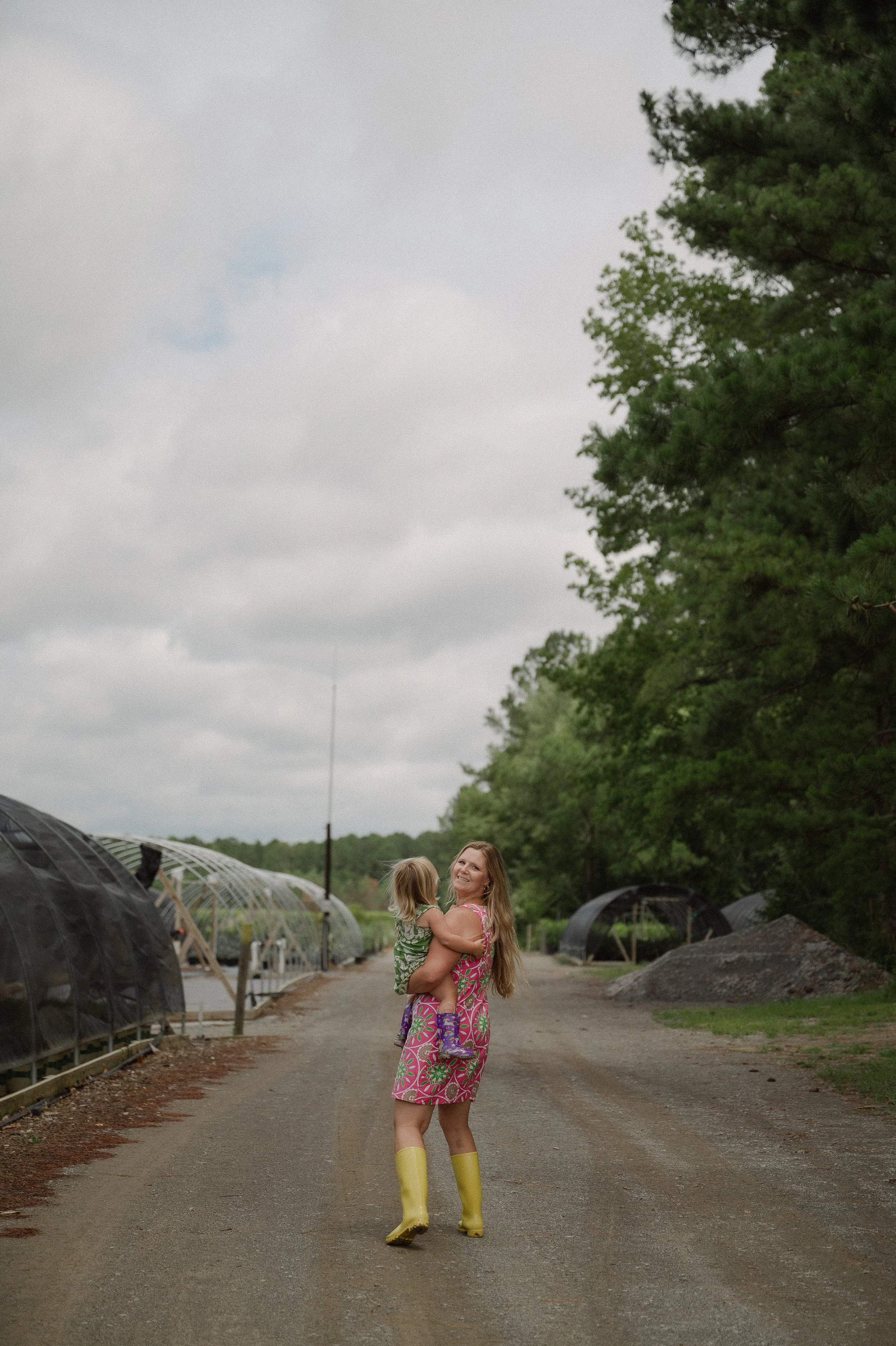 A woman wearing a colorful dress and yellow rain boots is smiling and holding a young girl who is wearing a green sweater and purple rain boots. They are walking on a dirt path in an outdoor setting with green trees and greenhouse tunnels on either side.