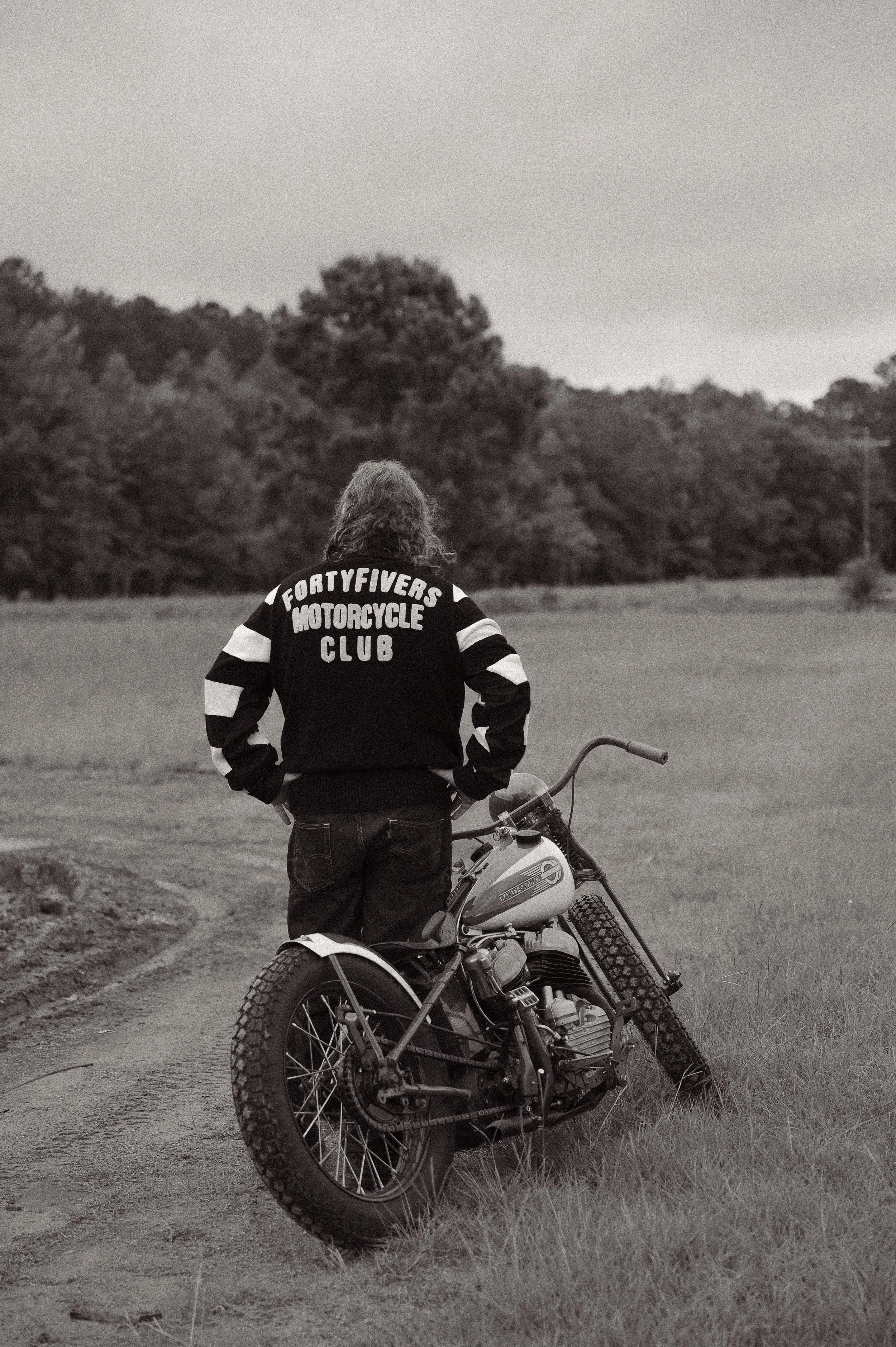 A person with wavy hair wearing a jacket with 'FortyFivers Motorcycle Club' on the back, standing outdoors with a vintage motorcycle on a dirt trail, surrounded by grass and trees in the background, in black and white.
