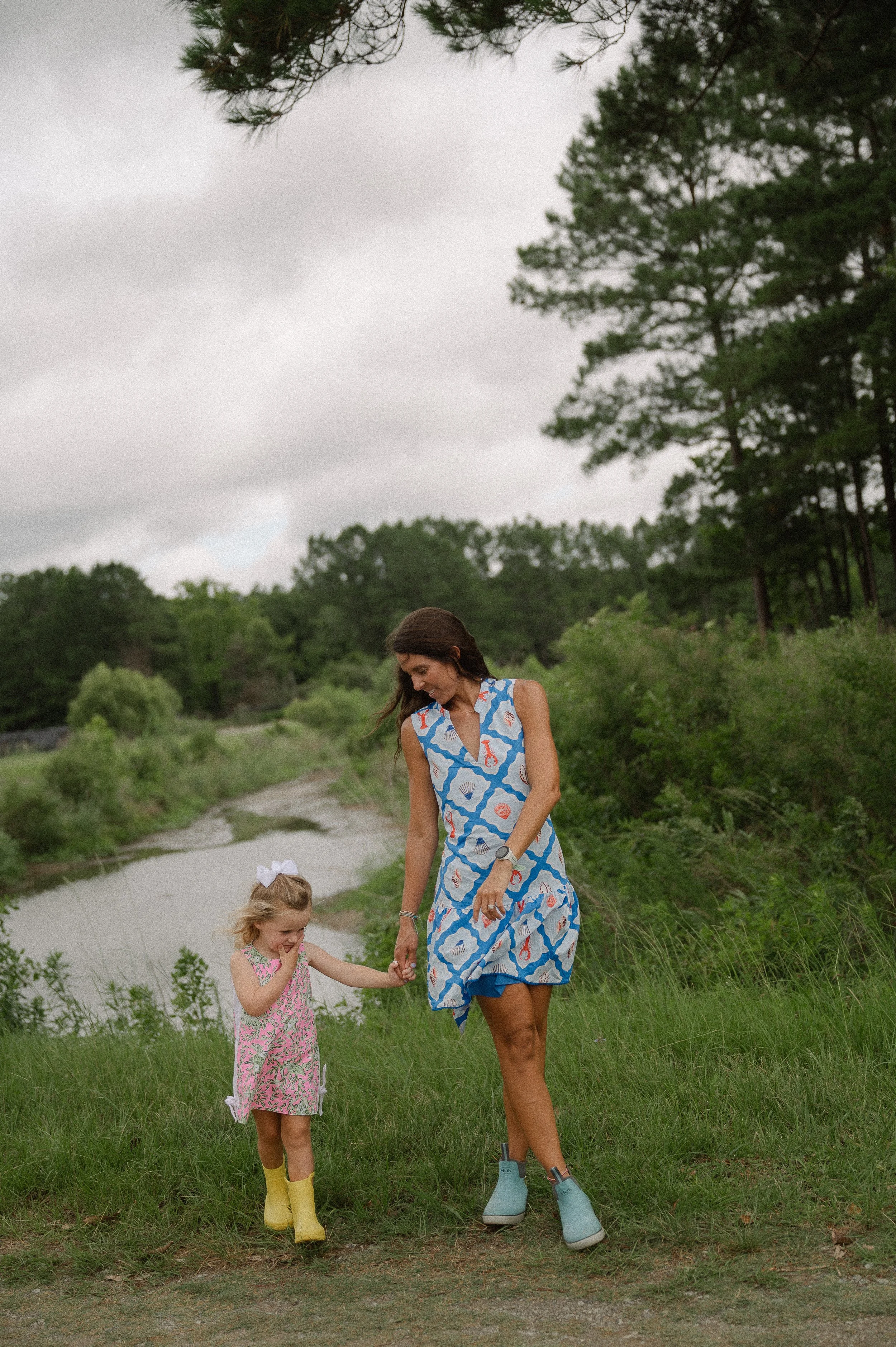 A woman and a young girl walking hand in hand along a grassy path near a river, surrounded by trees on a cloudy day.