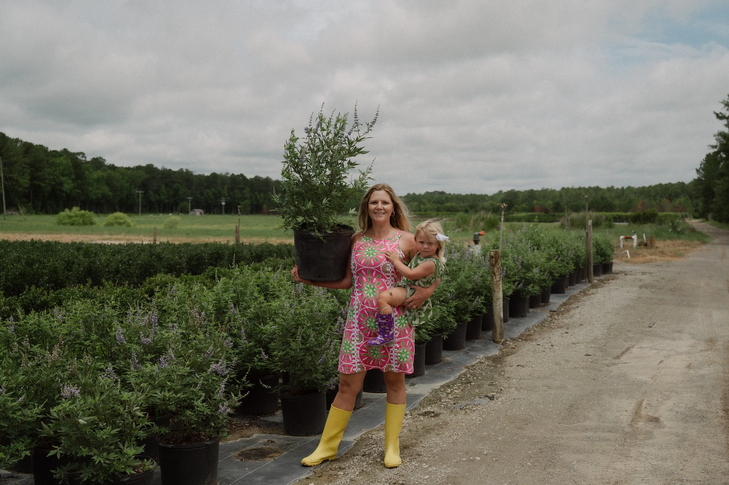 A woman and a young girl standing on a farm or nursery, with rows of potted plants and shrubs, holding a large potted plant, under a cloudy sky.