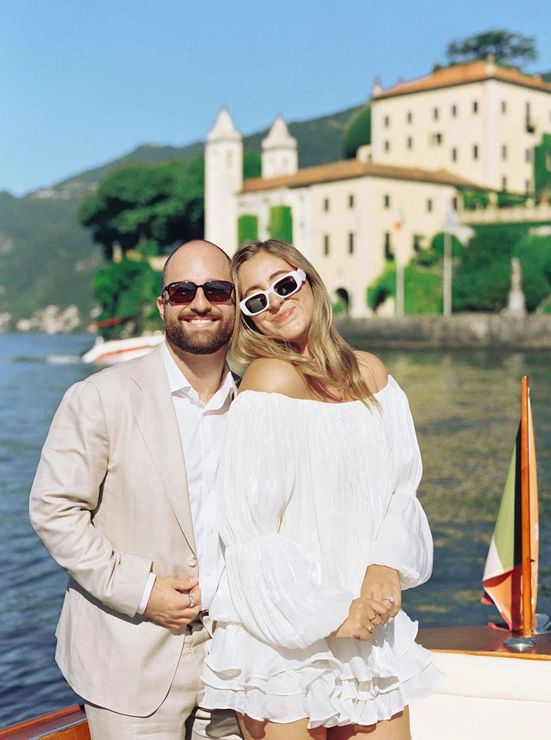 A smiling couple in white outfits and sunglasses on a boat with a European-style castle and green hills in the background.