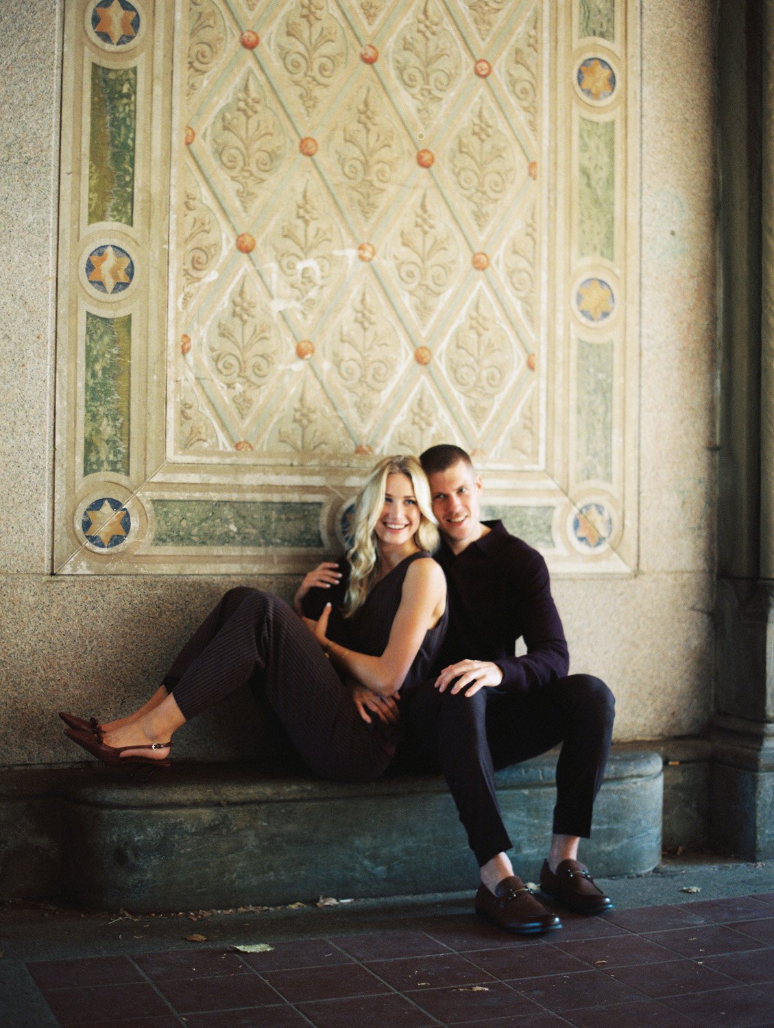 A young couple sitting on a bench in front of a decorative wall, smiling at the camera. The woman has blonde hair and is wearing a sleeveless black top and dark pants, while the man has short hair and is dressed in a dark shirt and pants.