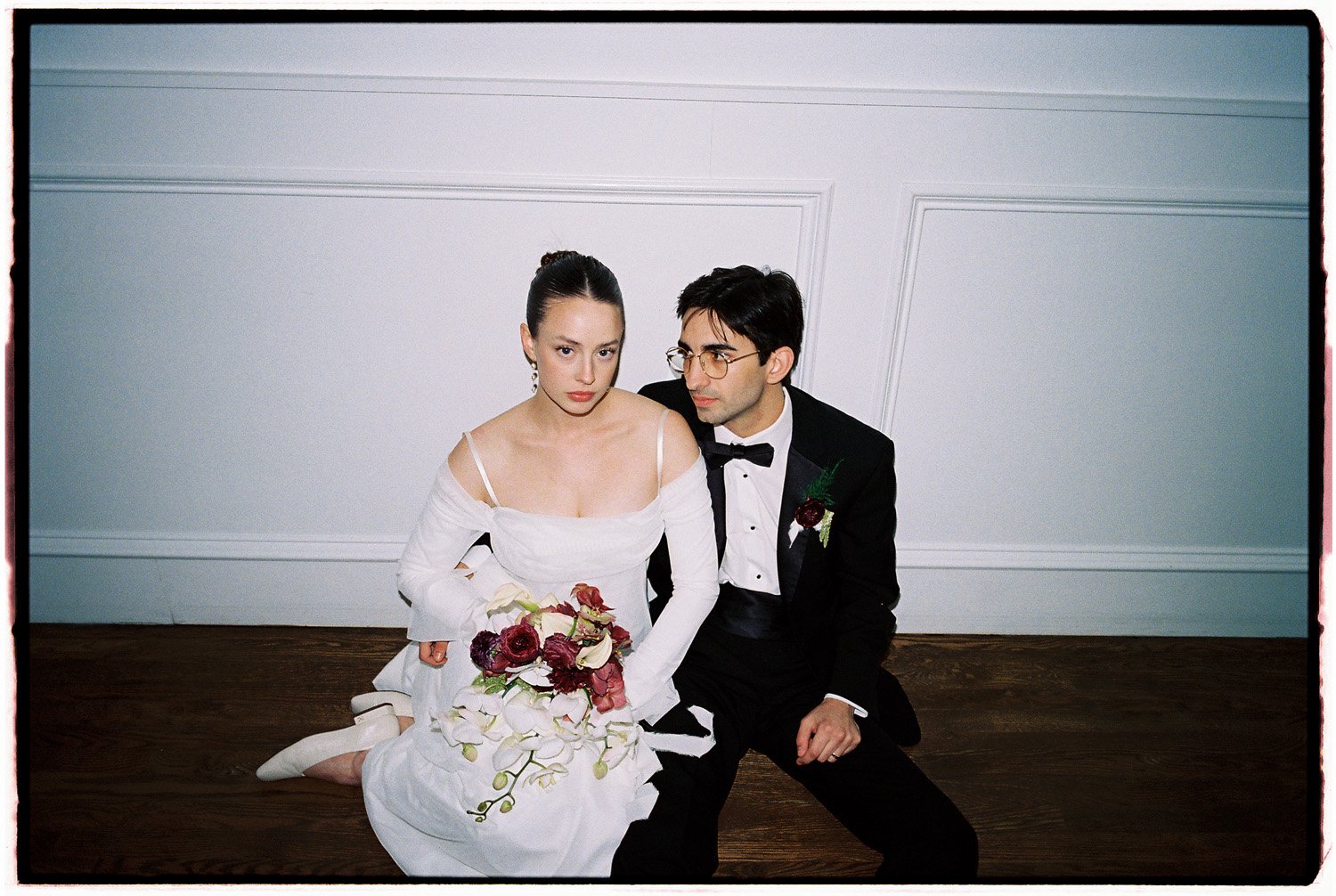 A man and woman dressed in wedding attire, sitting on the floor with a white wall background. The woman holds a bouquet of flowers and wears a white off-shoulder dress, while the man wears a black tuxedo with a bow tie and glasses.