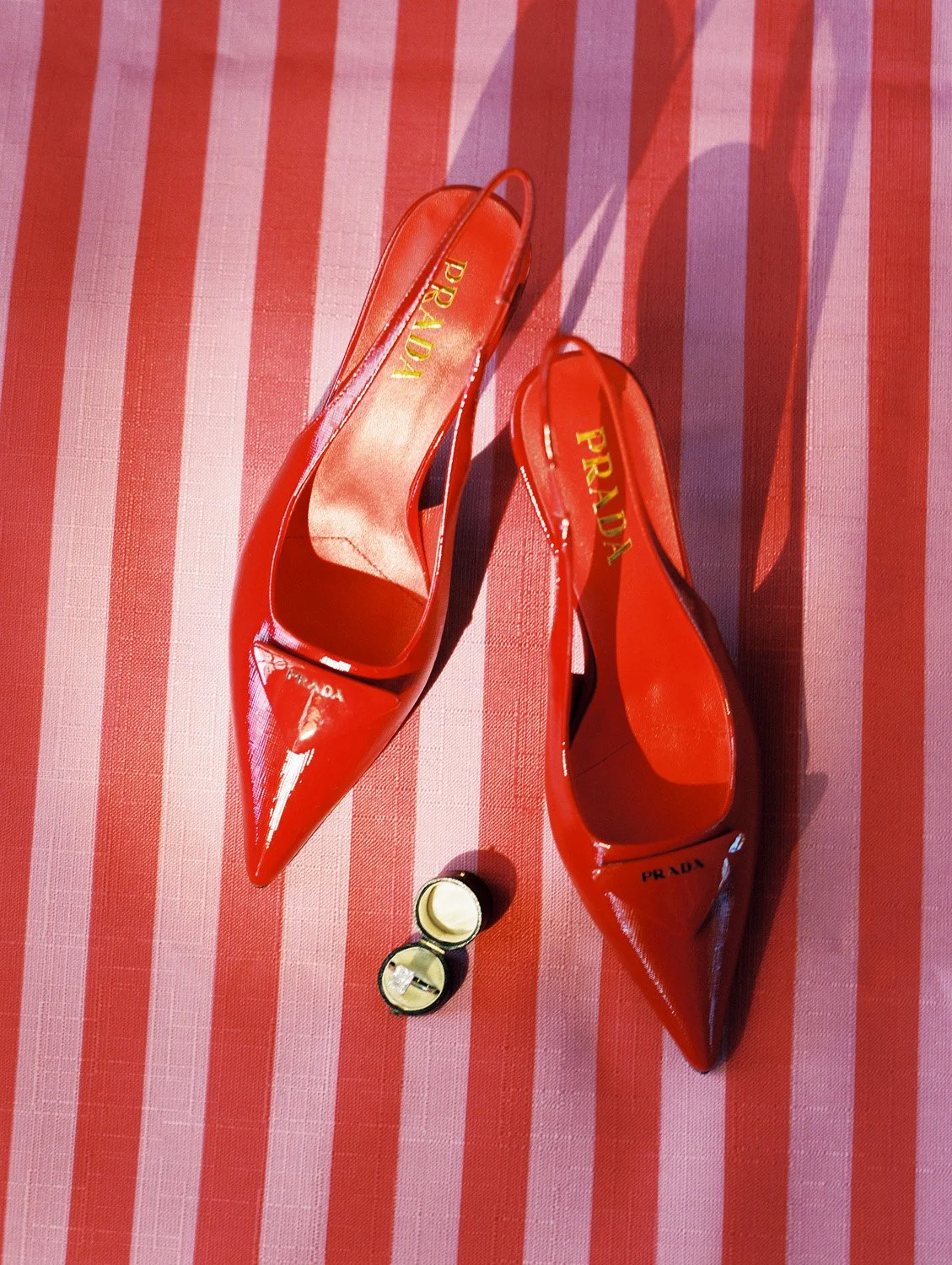 A pair of red Prada high-heeled shoes with pointed toes, purple insole, and gold logo, placed on a red and pink striped fabric. A ring with a yellow band and a large clear gemstone is also on the fabric.