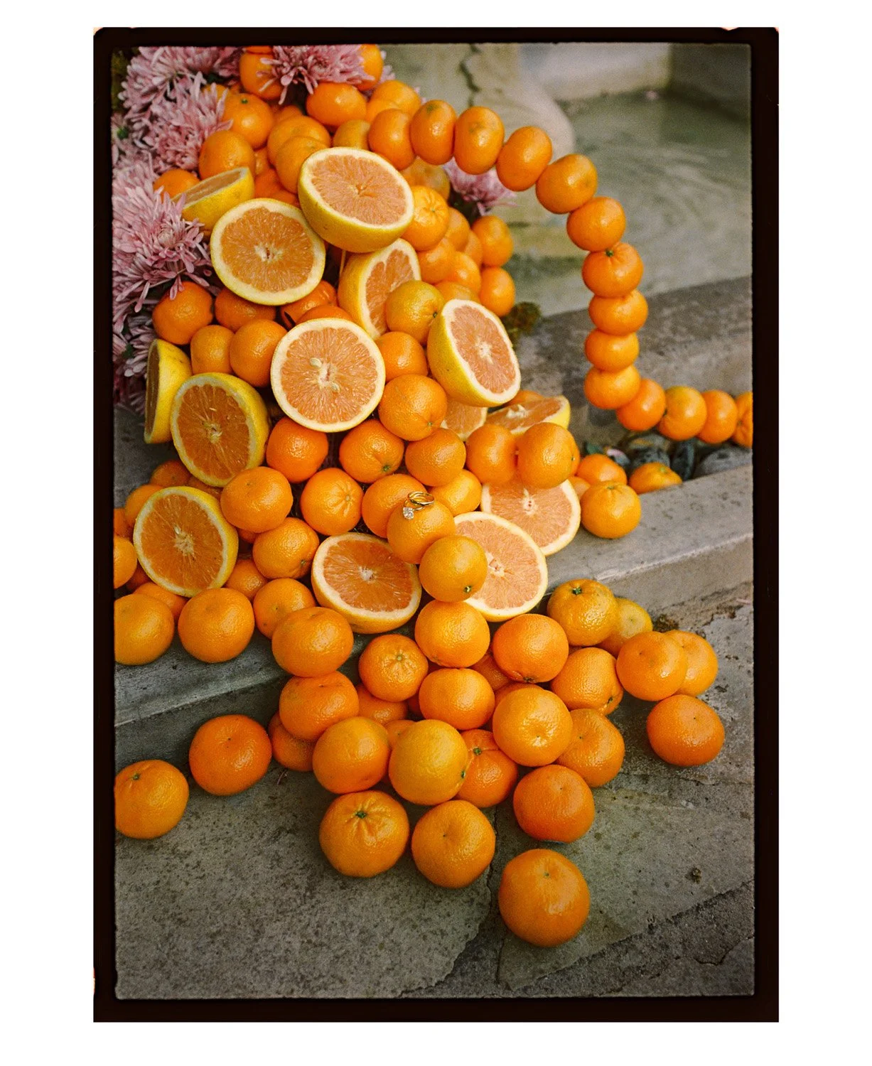 Orange citrus fruits, some sliced to reveal the interior, arranged on stone steps with pink flowers in the background.