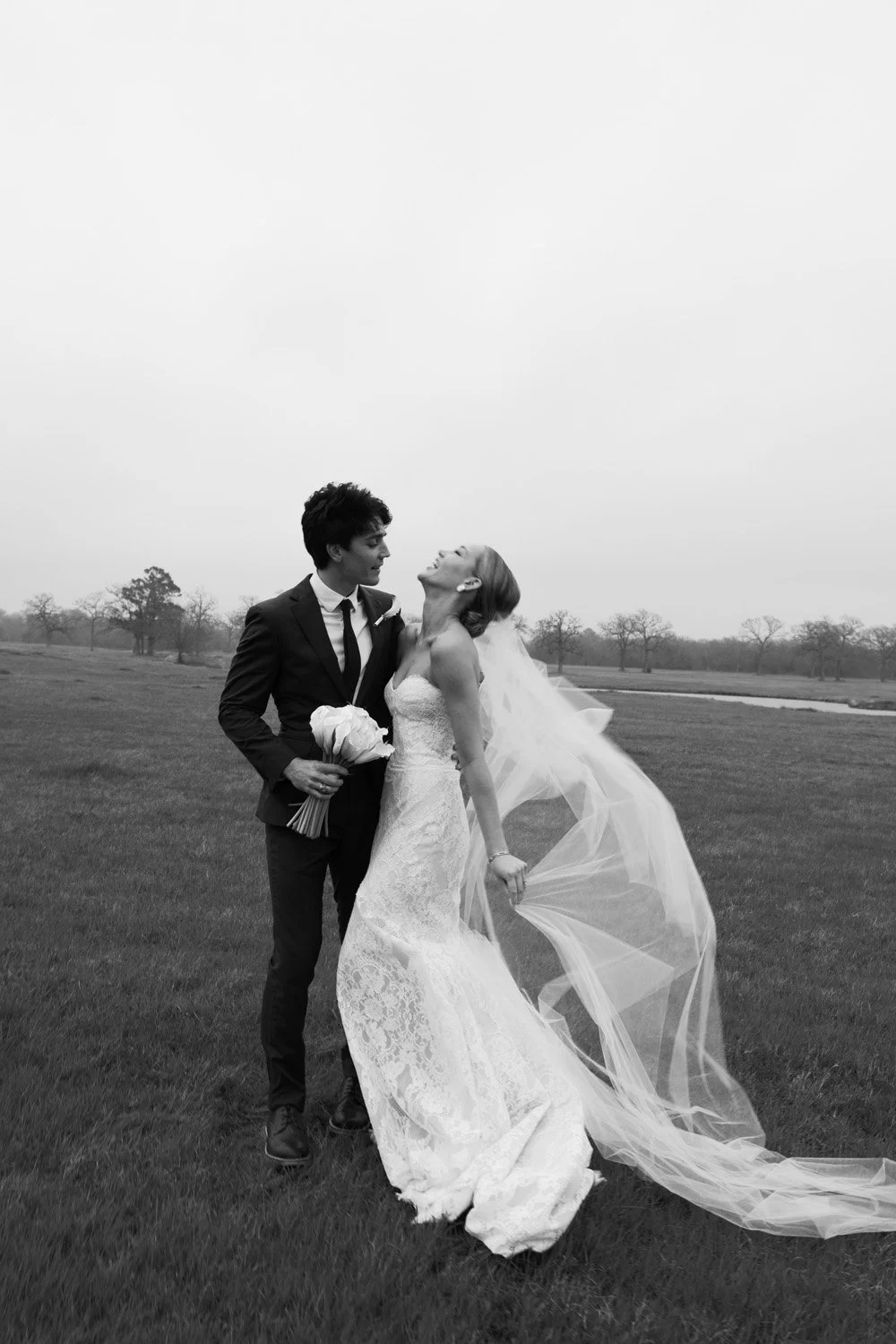 A black and white photo of a bride and groom on a grassy field, with the bride holding a bouquet and the groom looking at her.