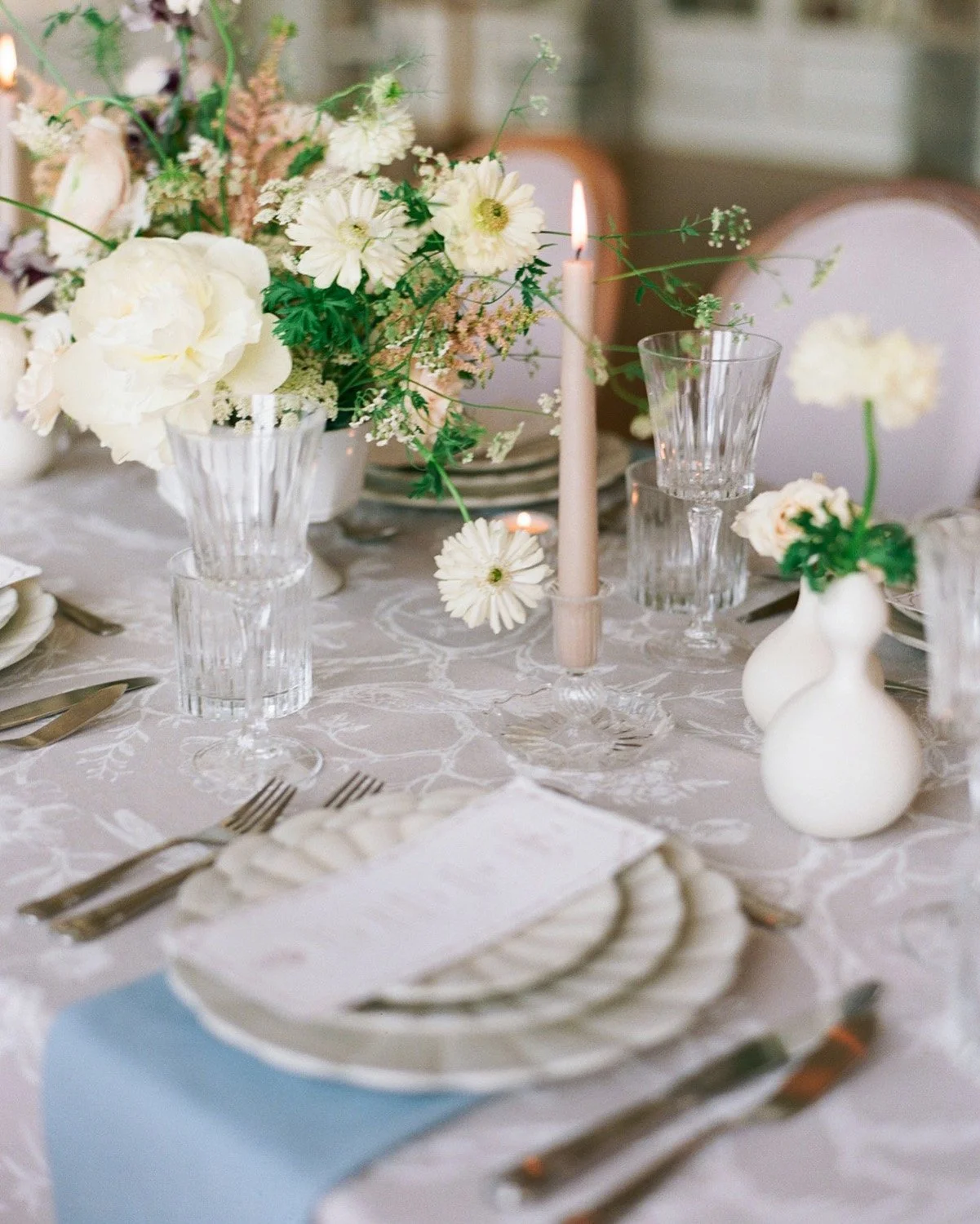 Elegant dining table setting with white floral centerpiece, white candles, clear glassware, and white patterned tablecloth.