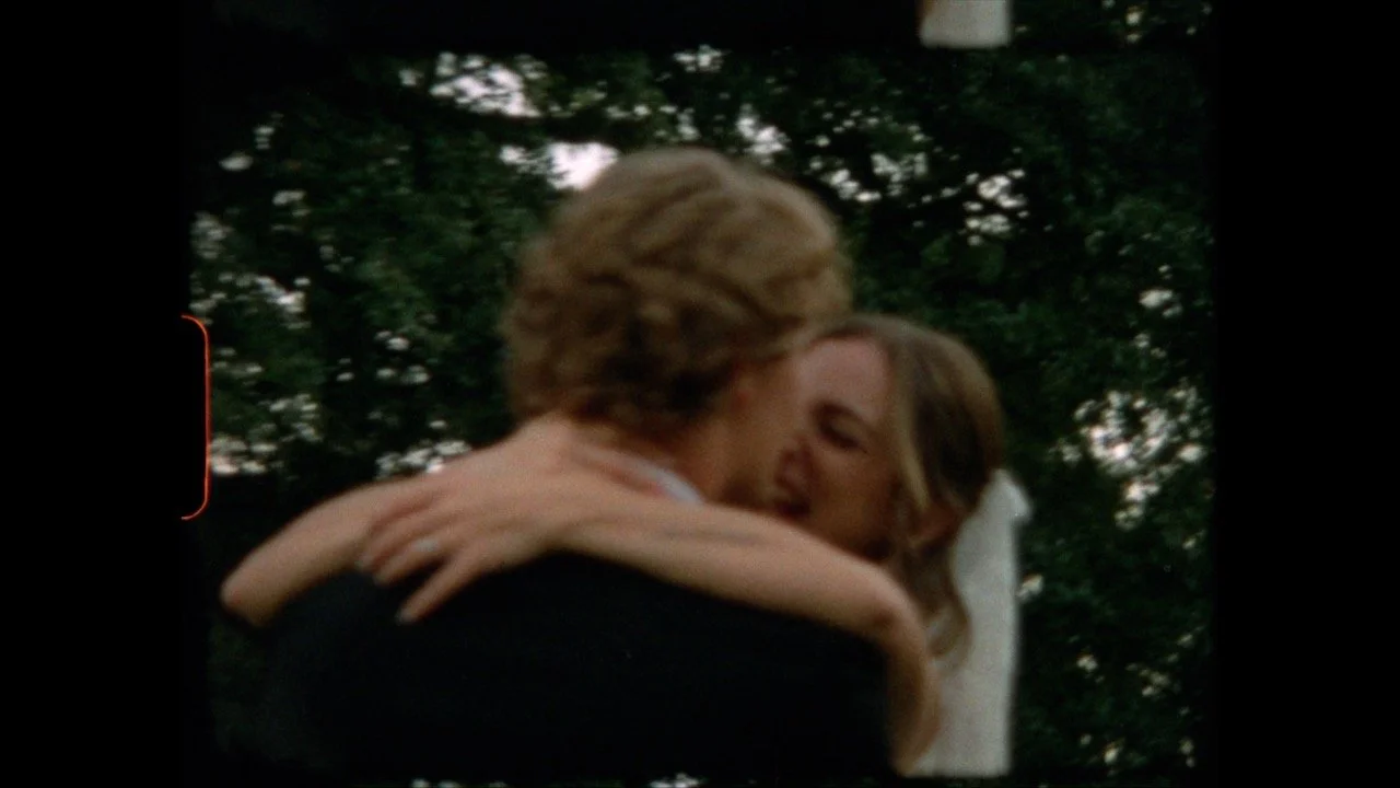 A man and woman embracing outdoors, with trees in the background.