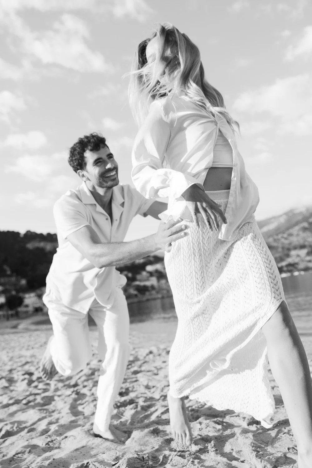 A joyful couple on the beach, with the man kneeling and smiling at the woman, who is standing and holding her dress as she laughs.