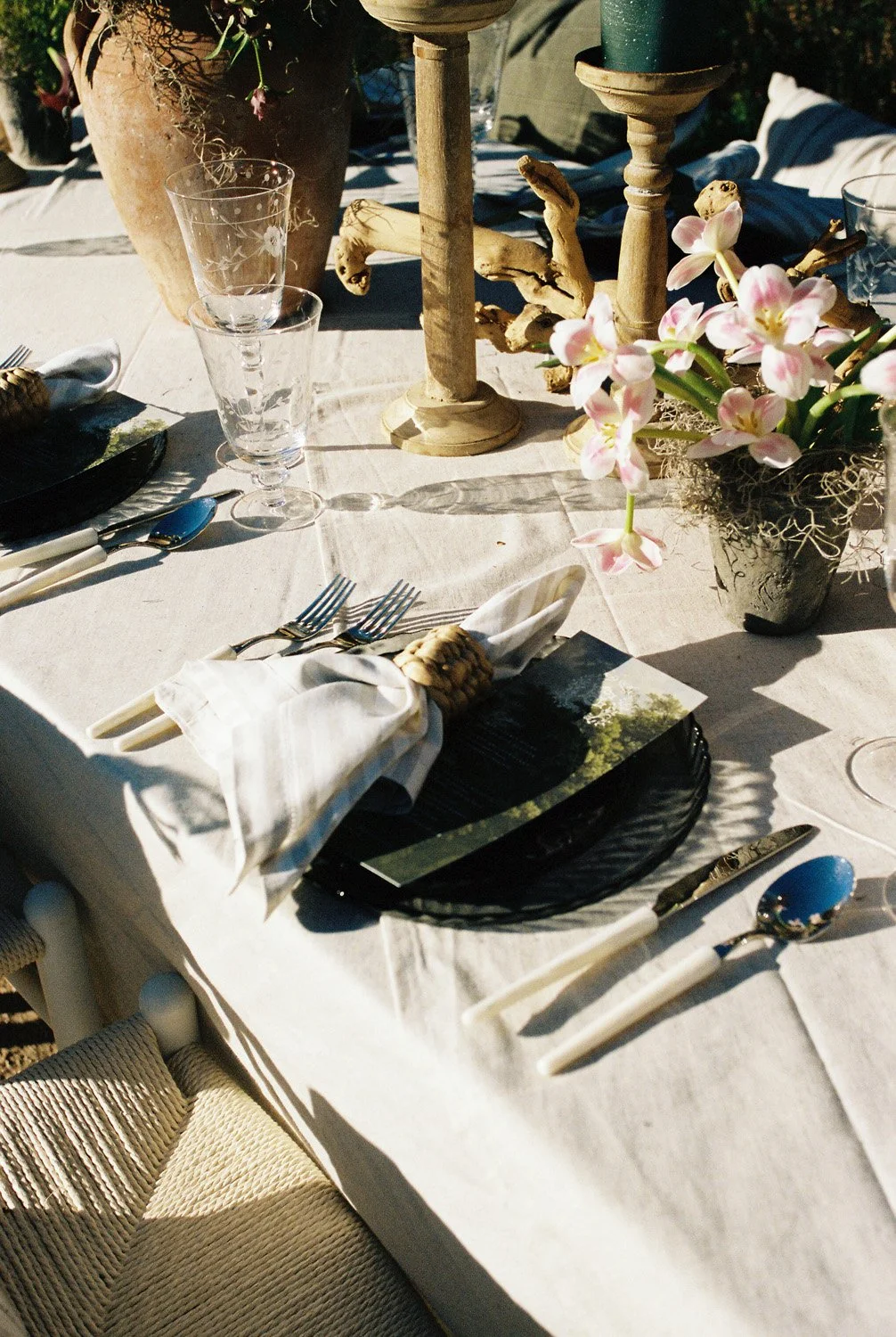 Elegant outdoor table setting with pink orchids in a black pot, two stacked wine glasses, silverware wrapped in a cloth napkin, black charger plate, and decorative candles with a large terracotta pot and driftwood centerpiece.