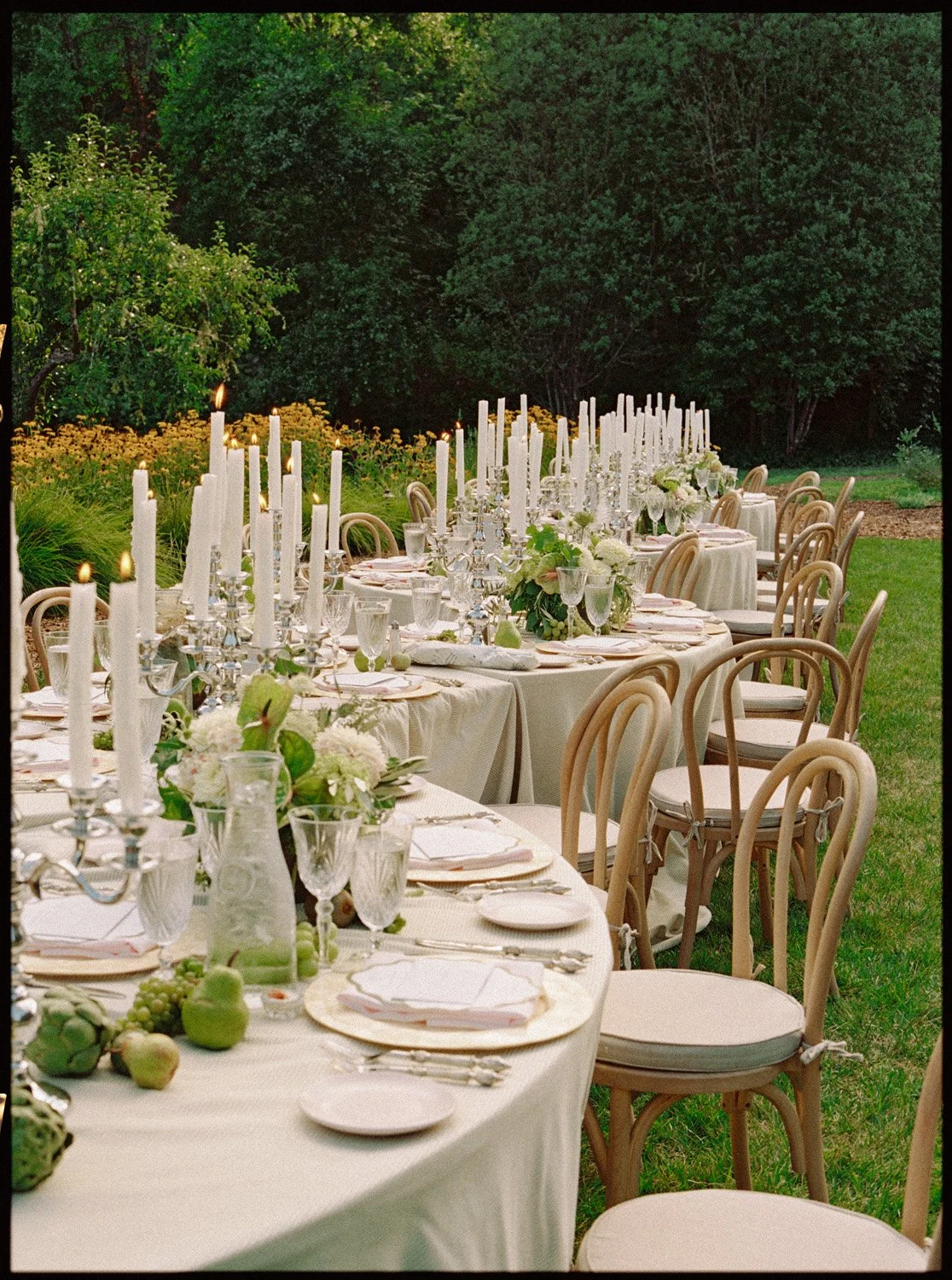 Outdoor banquet table set for a formal event with white tablecloths, candleholders with lit candles, floral centerpieces, and place settings, positioned on a lawn with greenery and trees in the background.