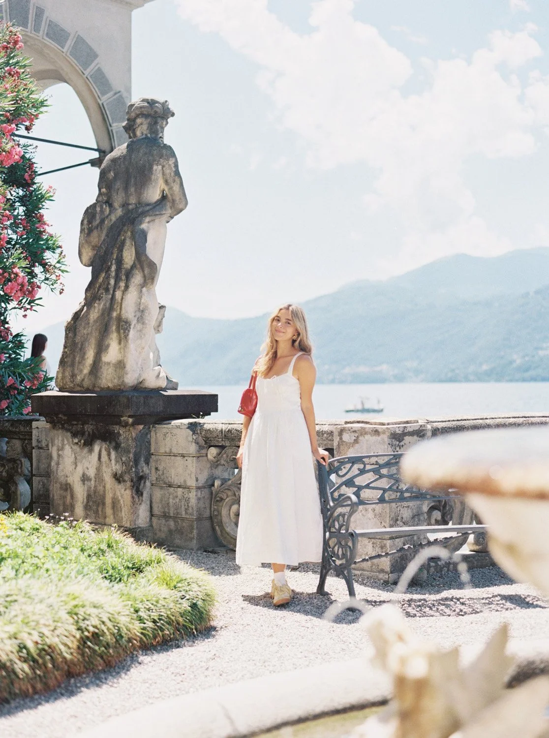 Young woman in a white dress standing on a terrace with a lake, mountains, a ship, a stone statue, and colorful flowers in the background.