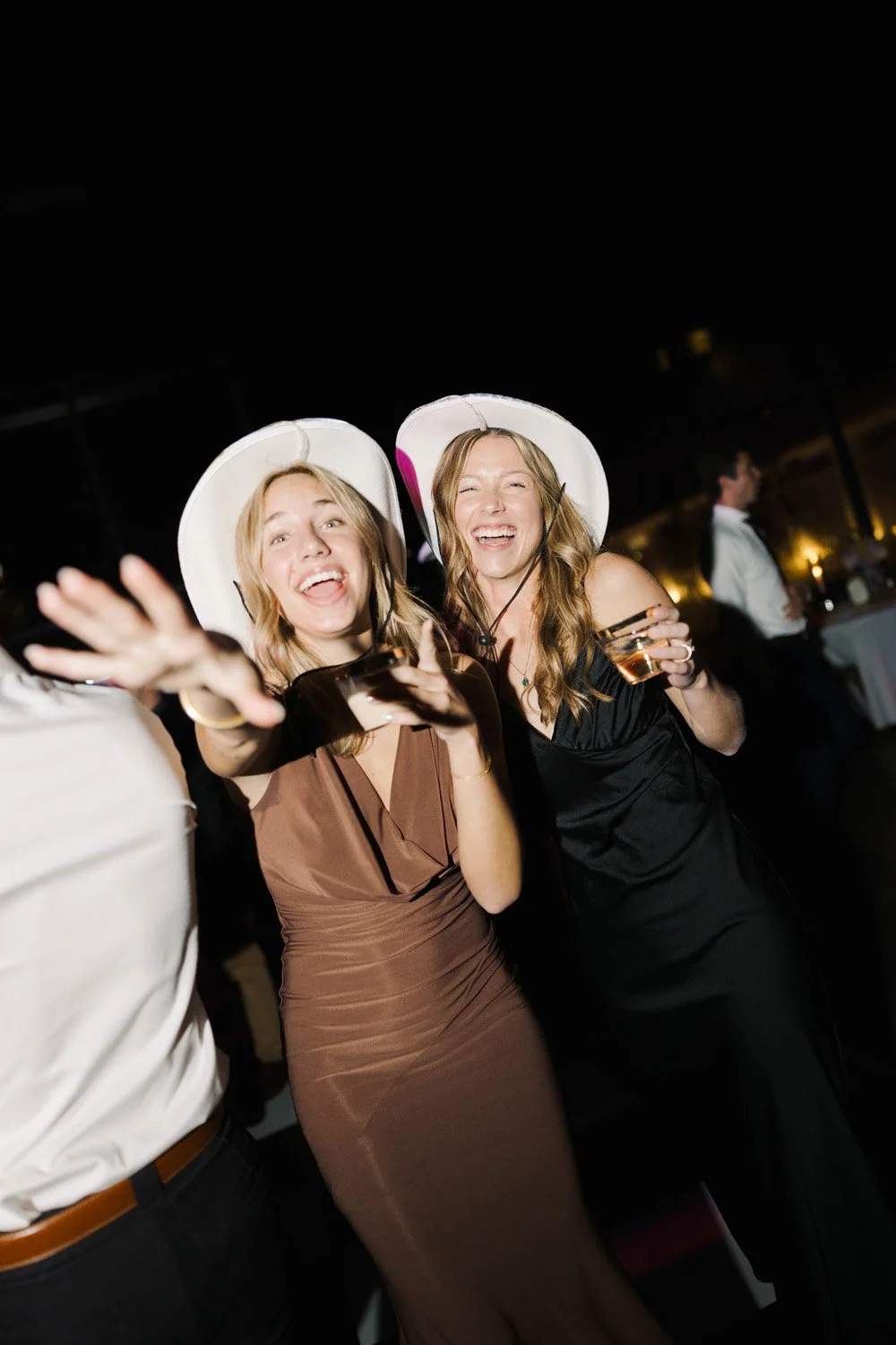 Two women wearing white party hats at a celebration, smiling, and holding drinks, with a dark background and some blurred figures in the background.